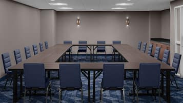 A conference room with a long table and blue chairs, equipped with overhead lighting and a kitchenette.