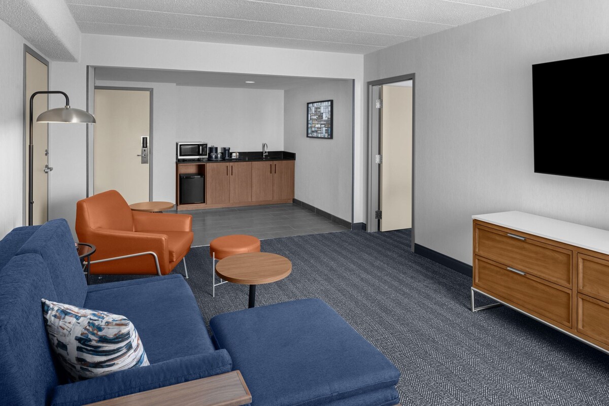 A modern hotel room with a blue sofa, orange armchair, small round table, and a wooden cabinet.