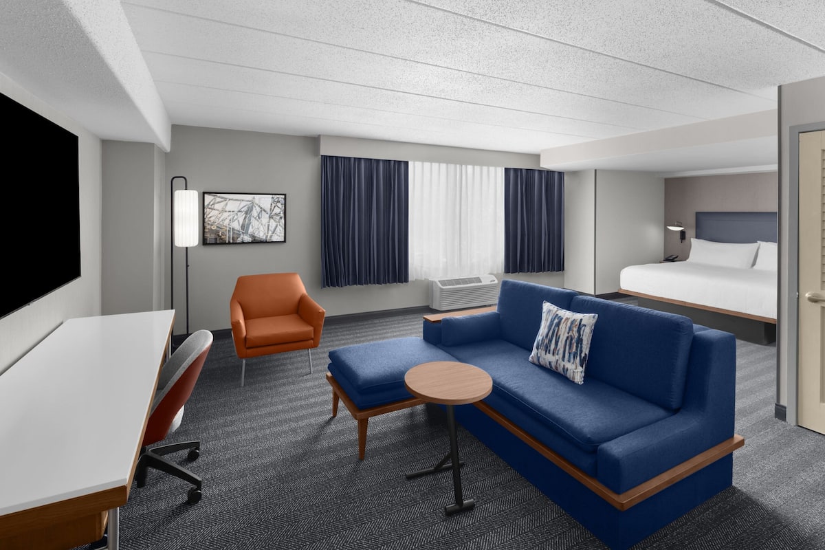 Een hotelkamer met een bed, een bank, een bureau, een oranje fauteuil, een ronde tafel en een groot raam met gordijnen.