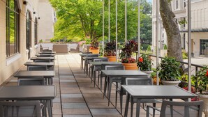 A covered outdoor seating area with tables and chairs, potted plants, and a view of a building in the background.