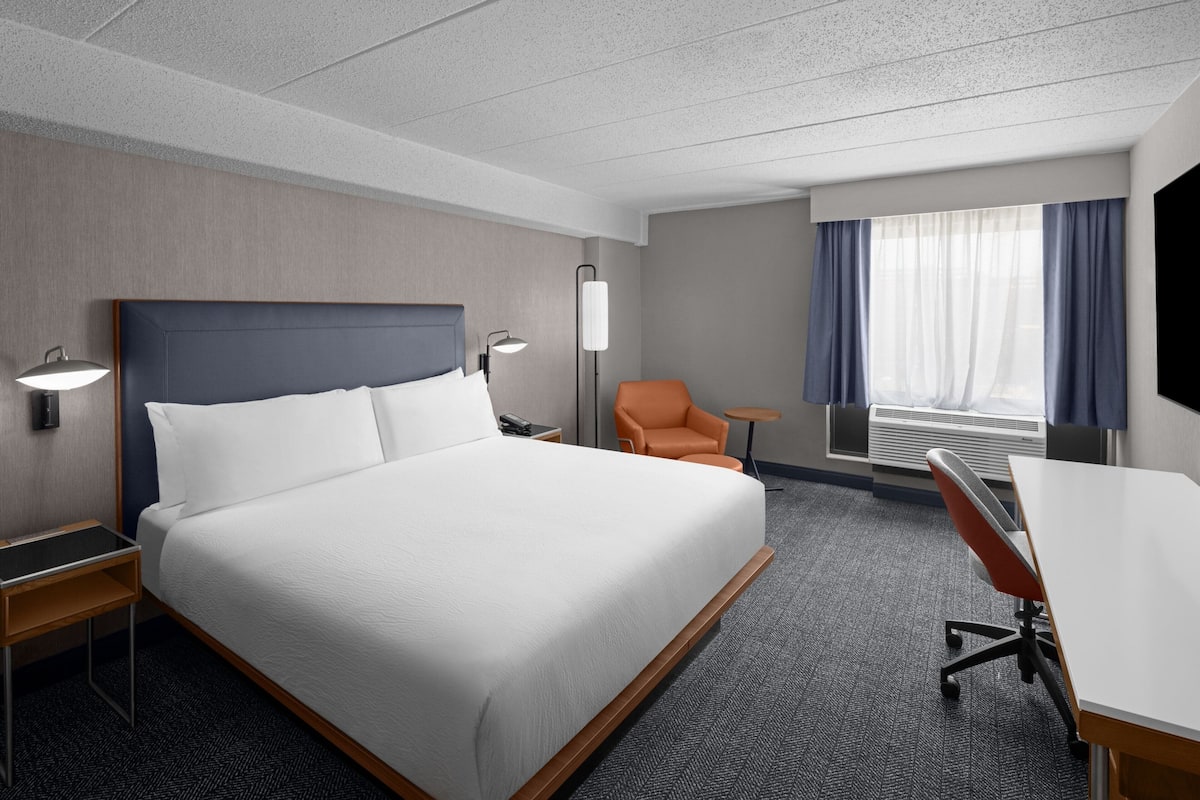 Een hotelkamer met een groot bed, een bureau, een oranje fauteuil, een lamp en een raam met gordijnen.