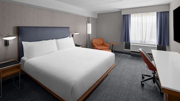 A hotel room with a large bed, a desk, an orange chair, a lamp, and a window with curtains.