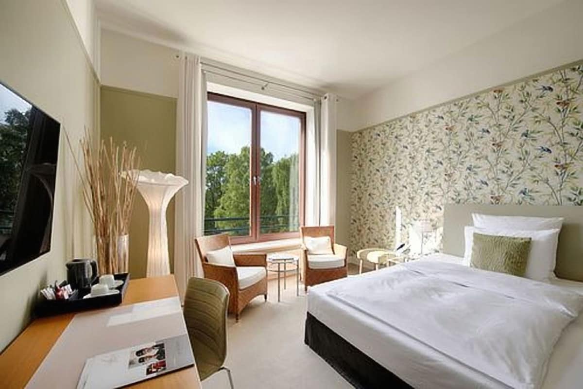 A hotel room with a bed, a desk, a chair, a small table, and a window with a view of trees.