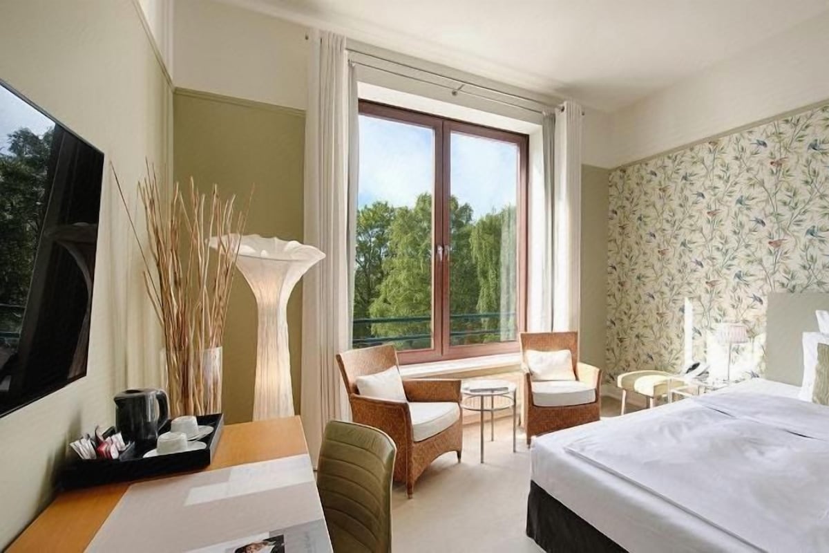 A hotel room with a bed, a desk, a chair, a small table, and a window with a view of trees.