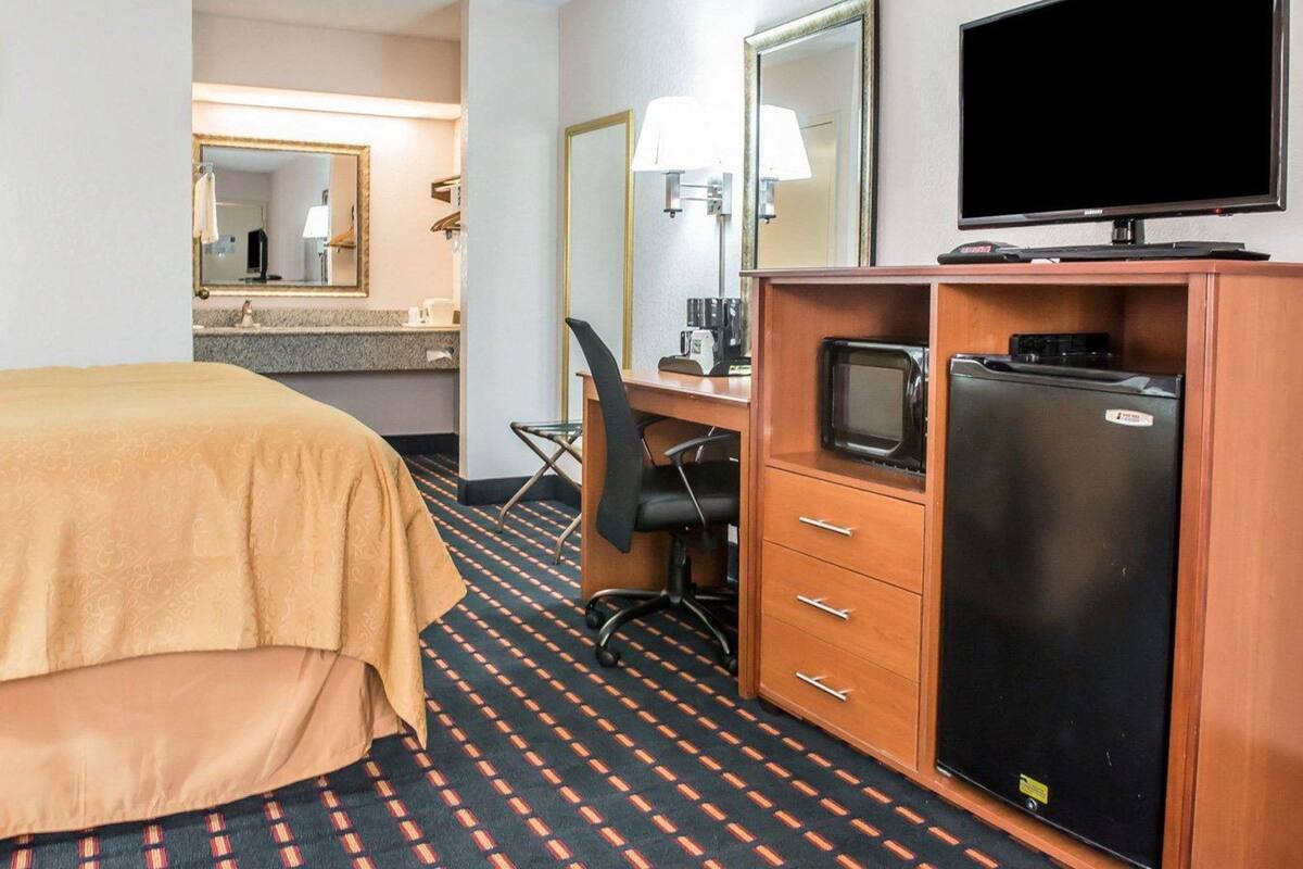 A hotel room with a bed, a desk with a computer, a refrigerator, a television, and a mirror.
