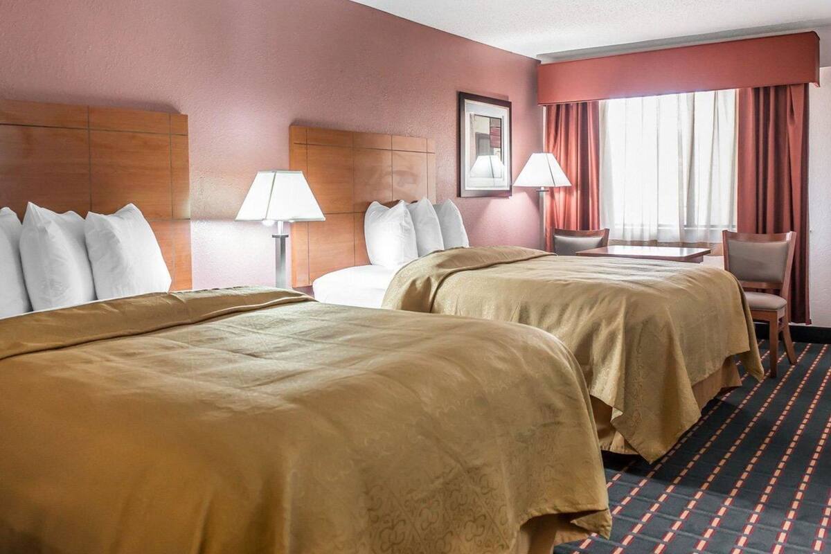 A hotel room with two beds, a desk, a chair, a lamp, a framed picture, and a window with curtains.