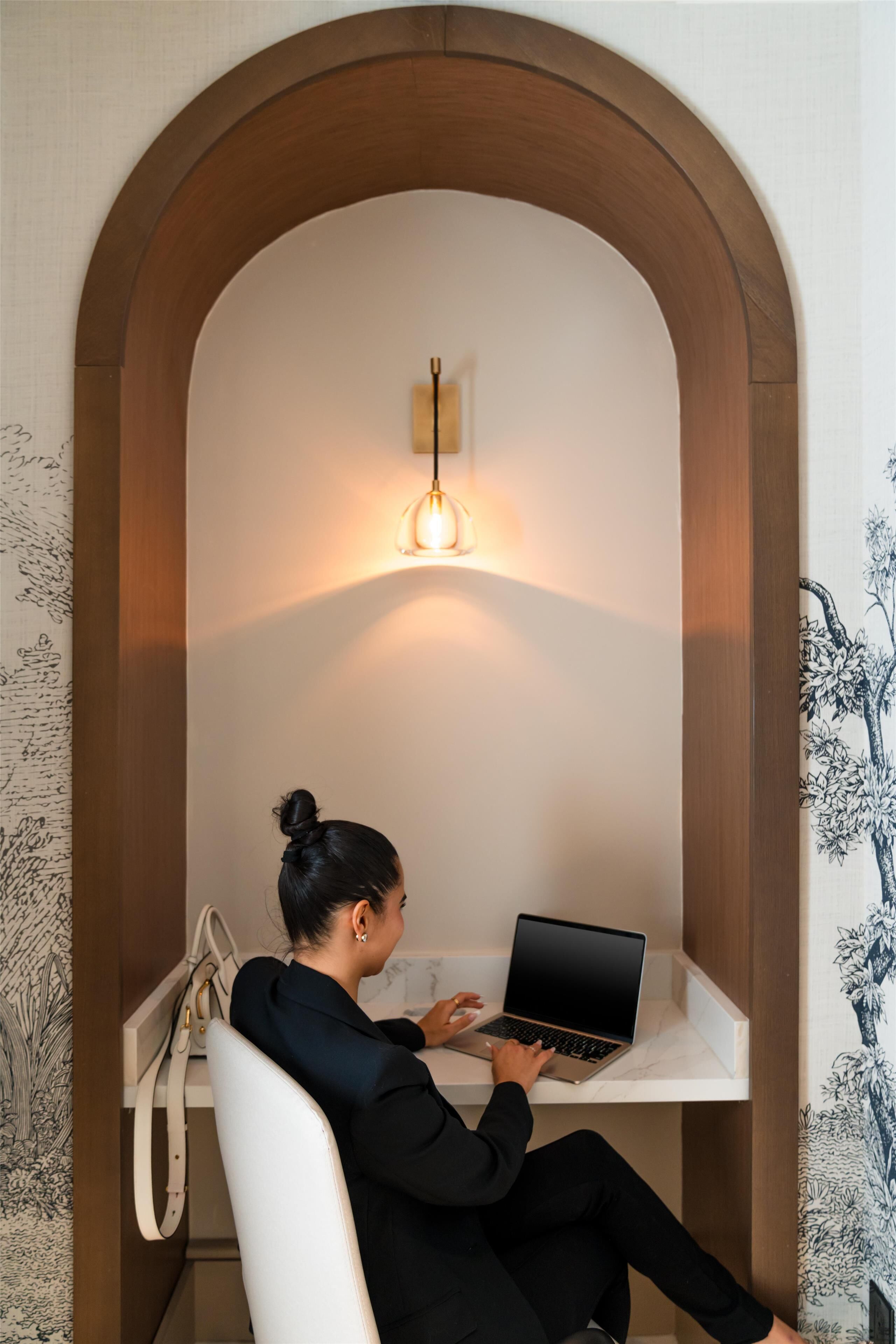 Une femme est assise à un bureau, devant un ordinateur portable, dans une pièce dotée d’une arche en bois et d’un luminaire fixé au mur.