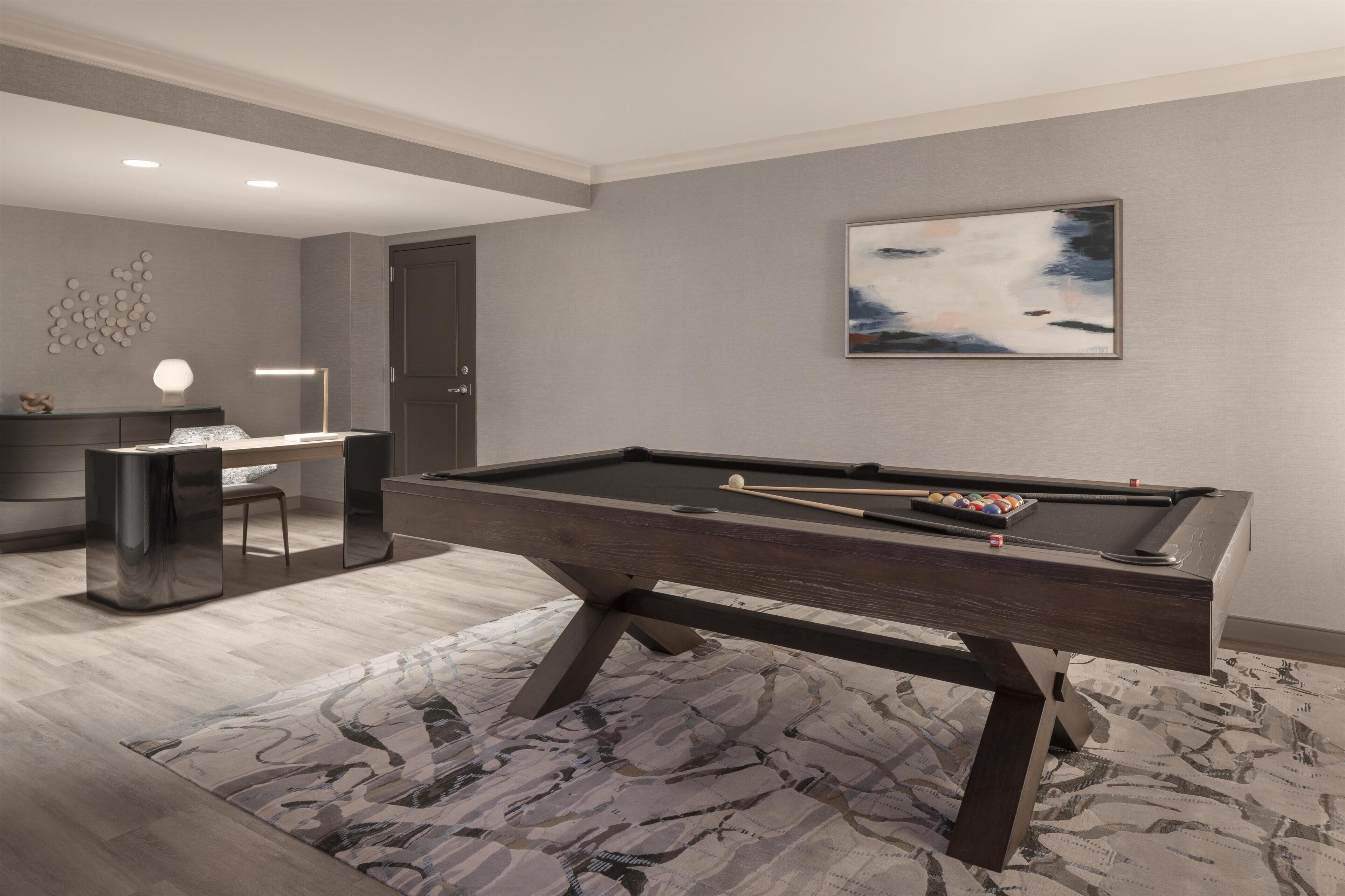 A modern pool table in a room with a neutral color scheme, a desk, and a framed abstract painting on the wall.