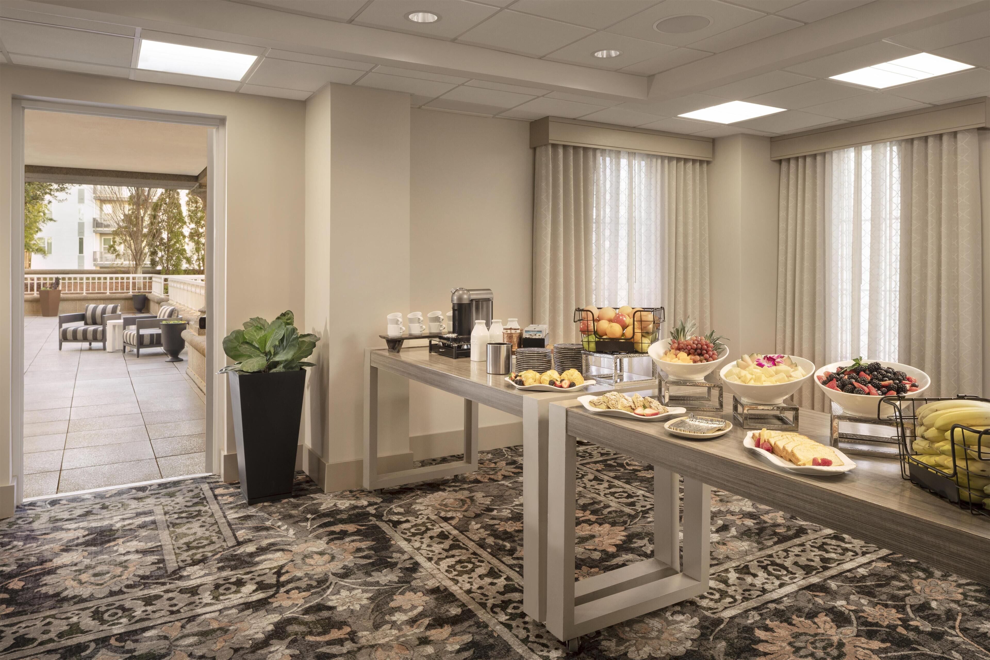 A buffet setup with various food items and beverages, a potted plant, and a view of an outdoor seating area.