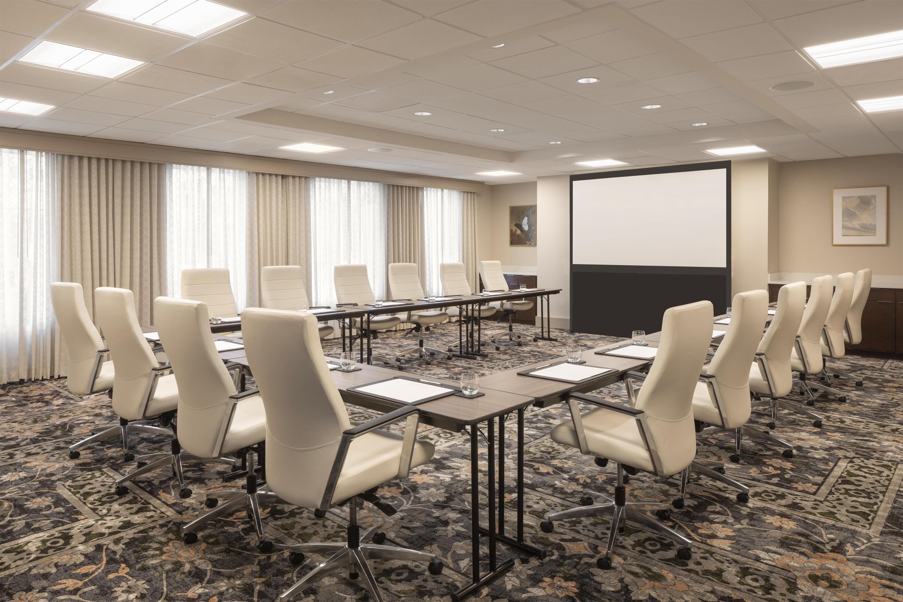 A conference room with a long table, multiple chairs, a projector screen, and a carpeted floor.