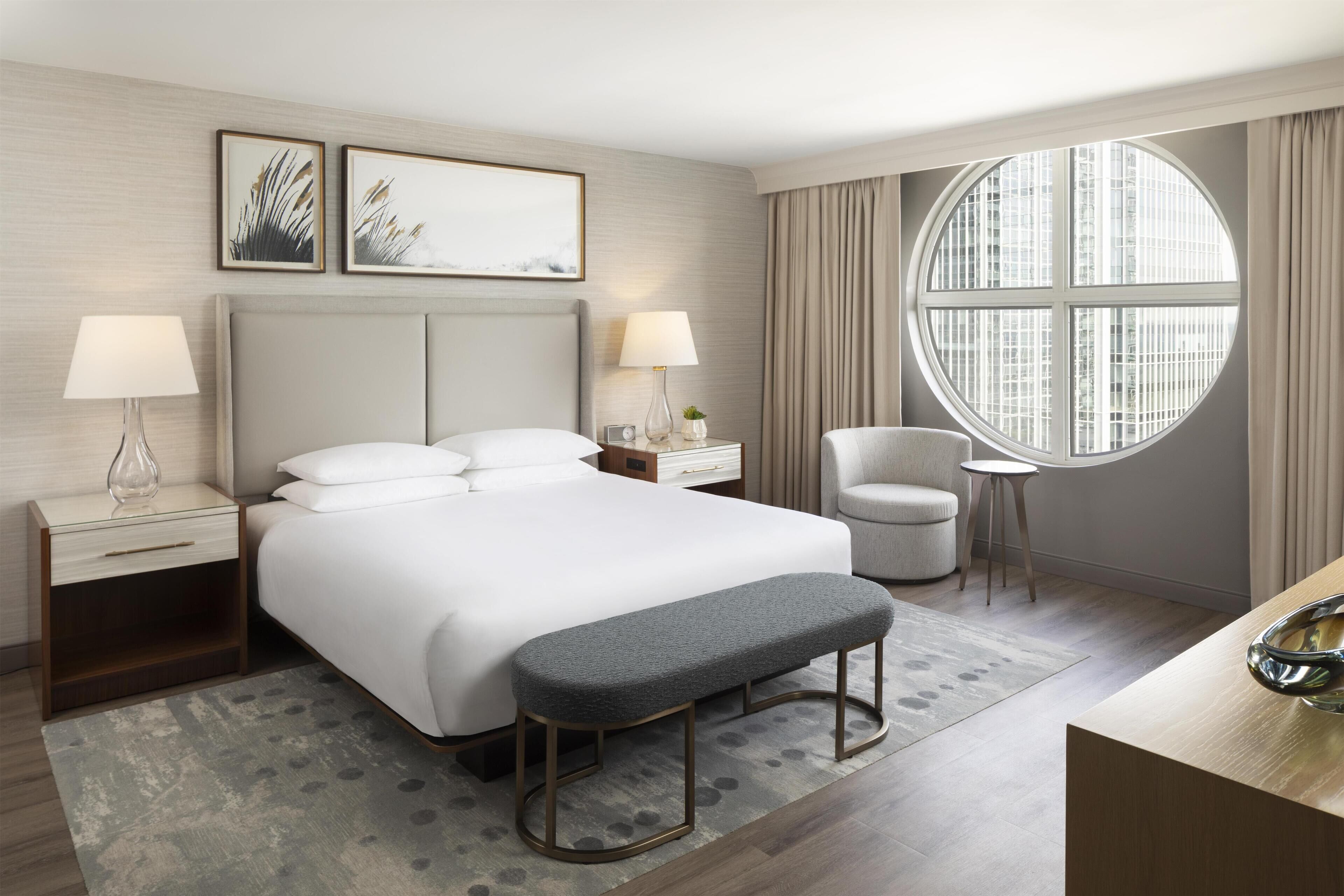A modern hotel room with a large bed, bedside tables with lamps, a round window, and a small round table.