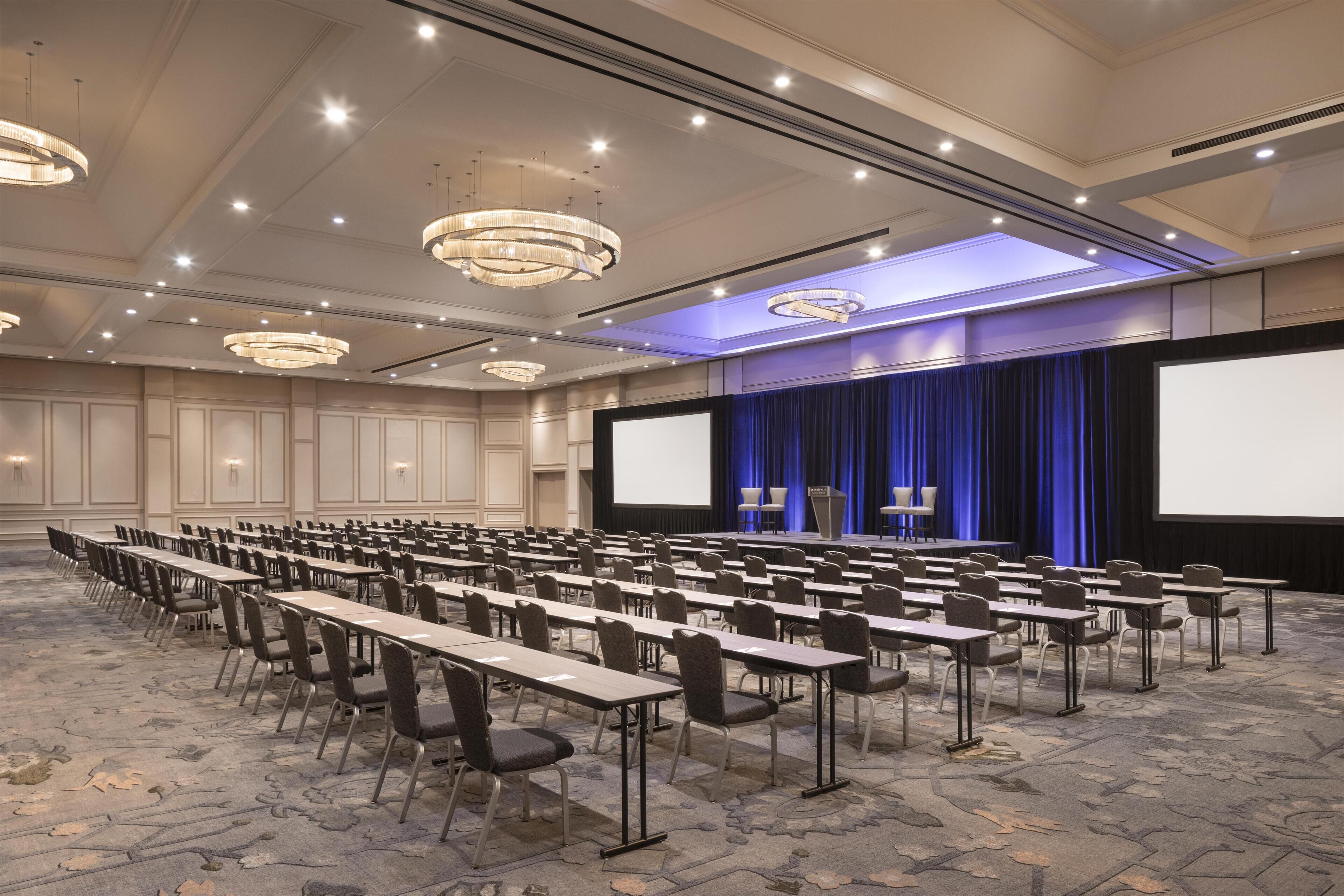 Un salón de conferencias con filas de mesas y sillas, una pantalla grande y un escenario con cortinas.