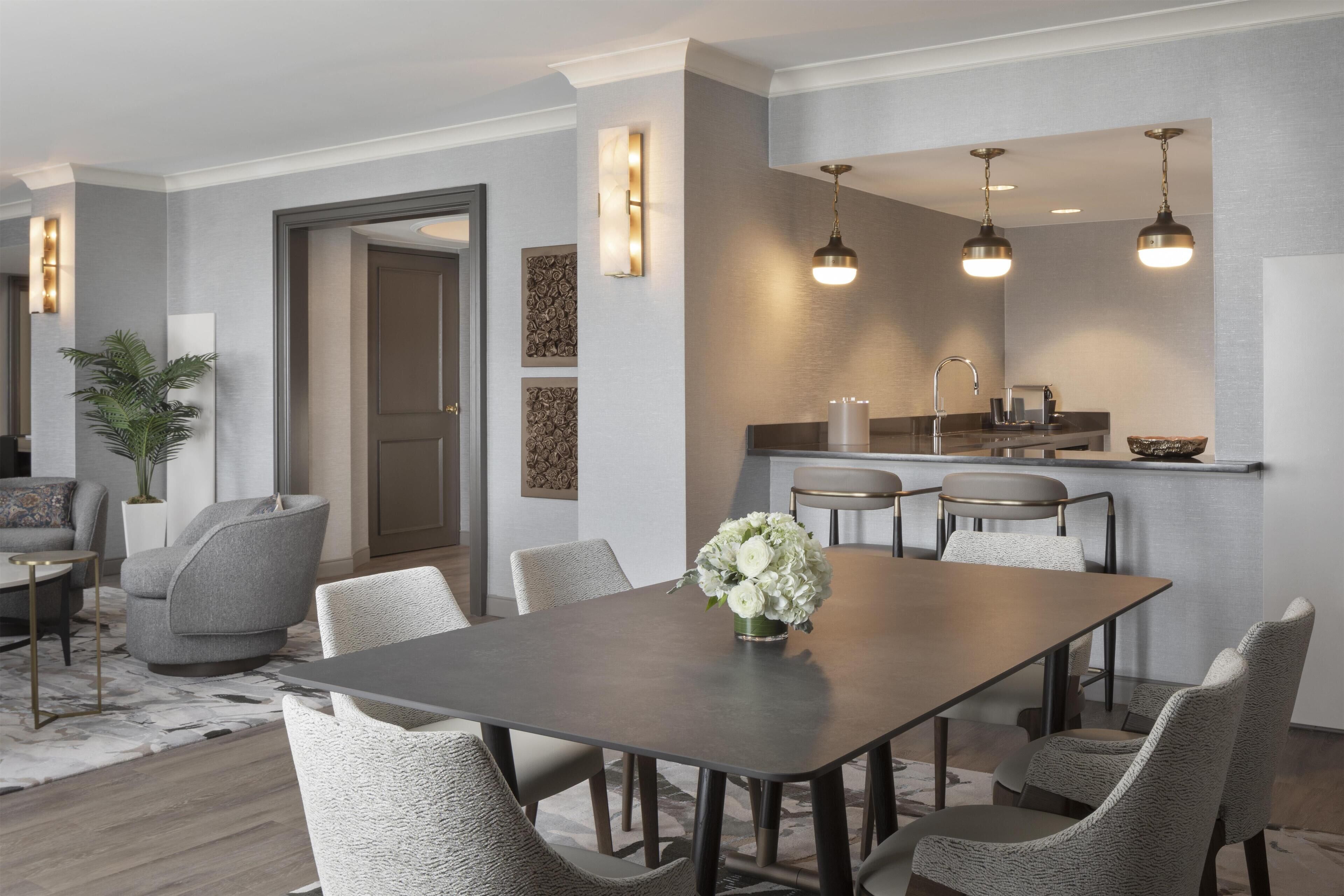 A modern dining area with a rectangular table, six chairs, and a vase of flowers. A kitchen with a sink and countertop is visible in the background.