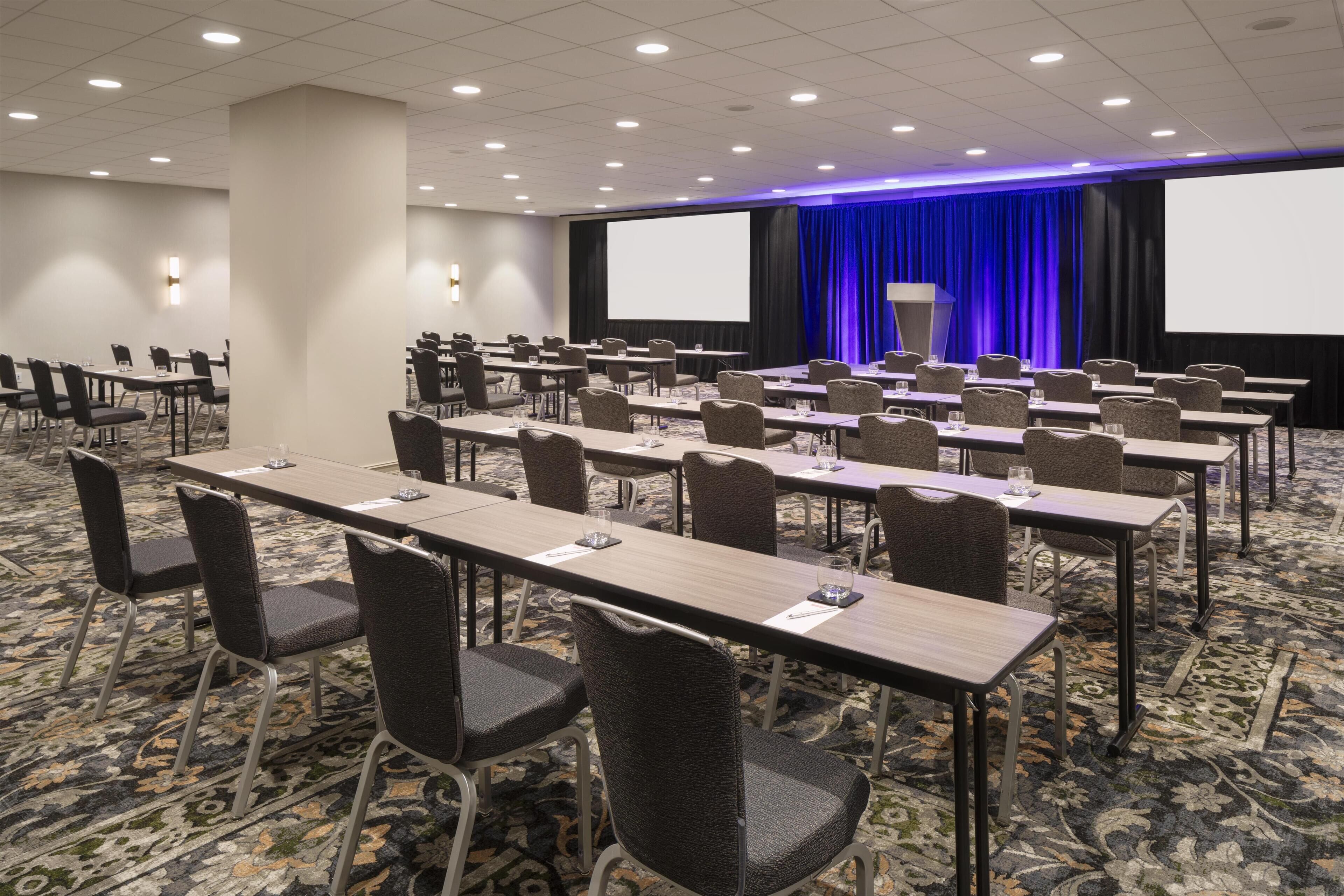 A conference room with rows of tables and chairs, a large screen, and a blue-lit stage area.