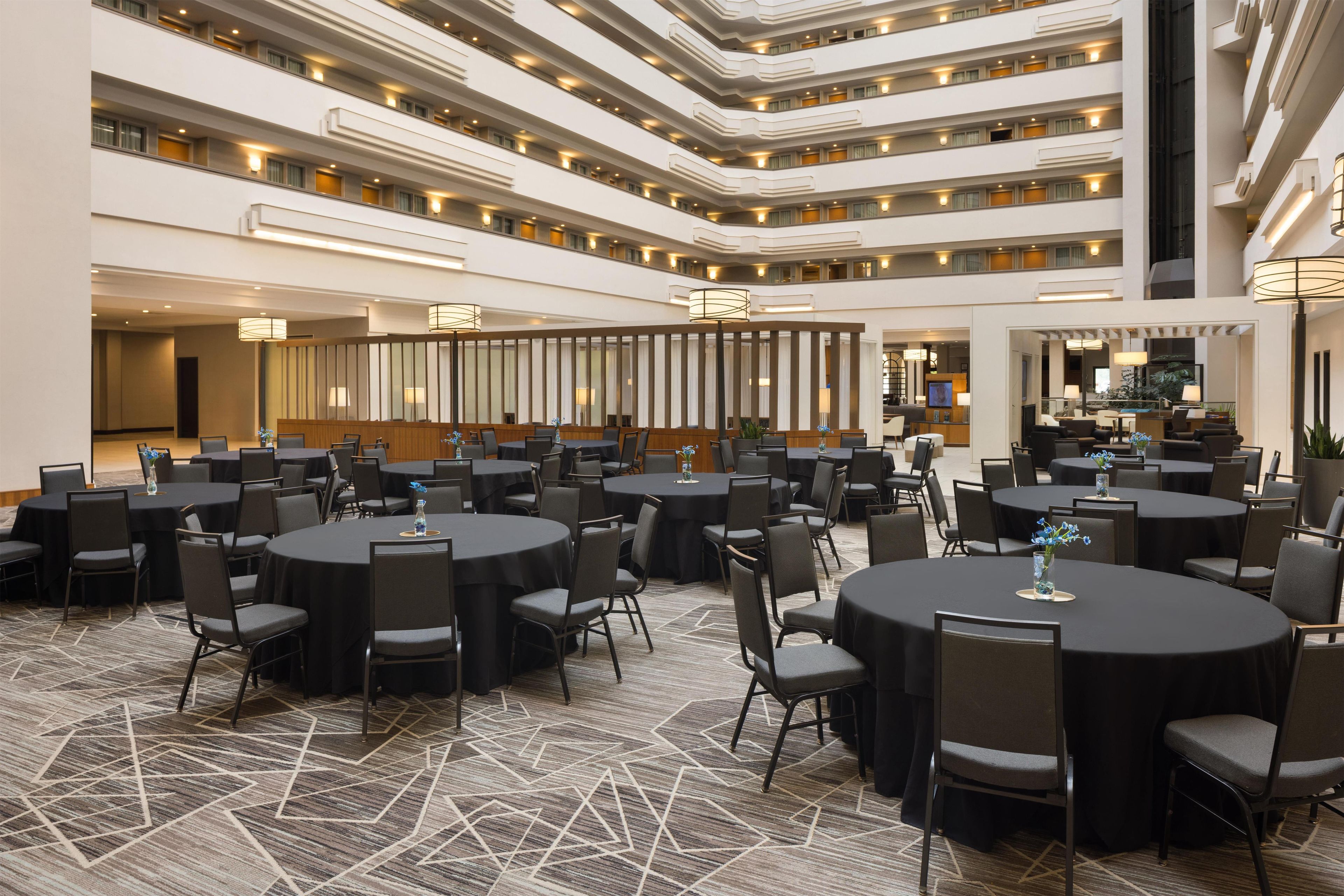Un hall d’hôtel à plusieurs niveaux, avec des tables et des chaises rondes, un espace de réception central et un escalier.