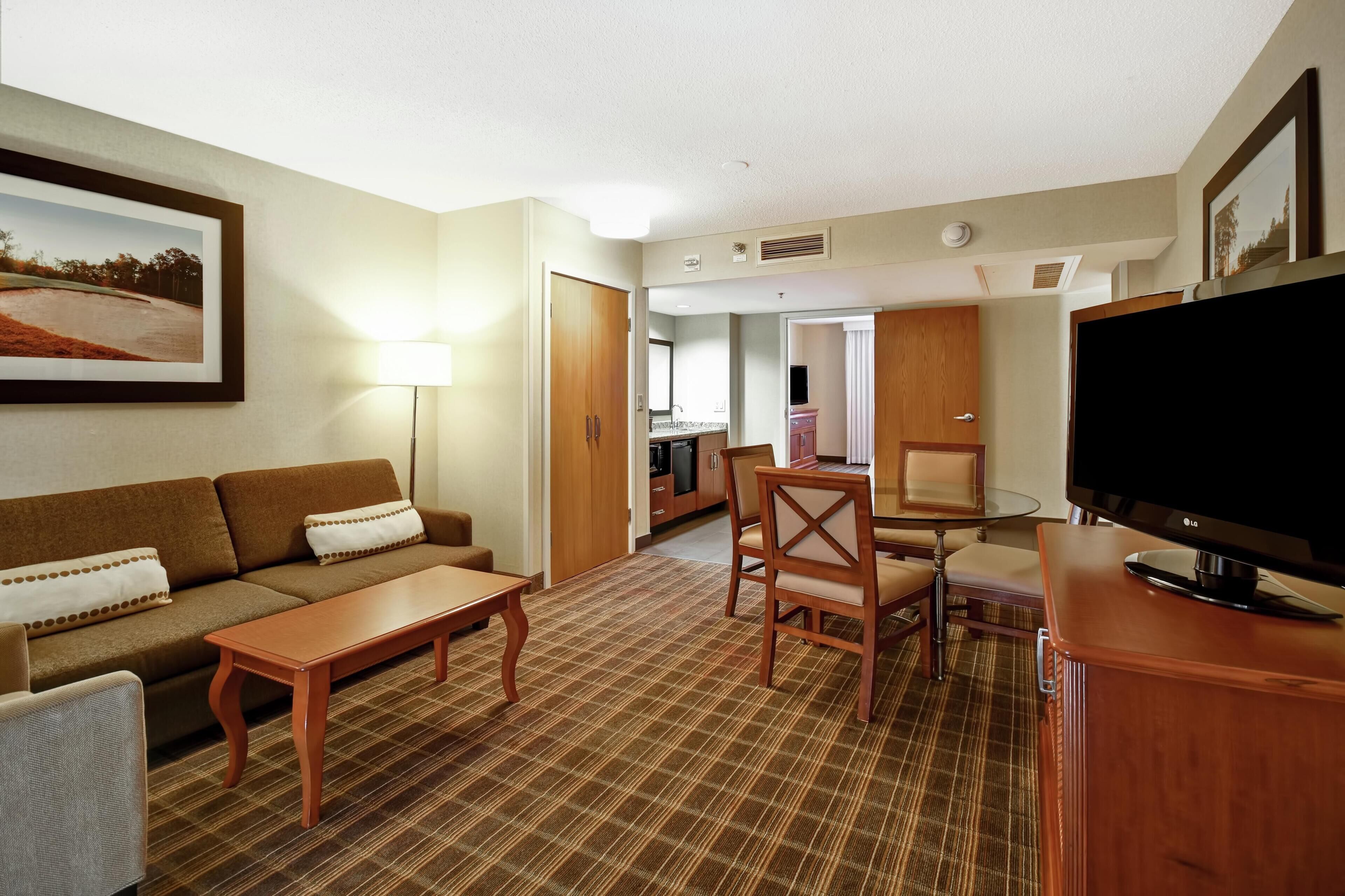 A hotel room with a sofa, a dining table, a TV, and a kitchen area in the background.