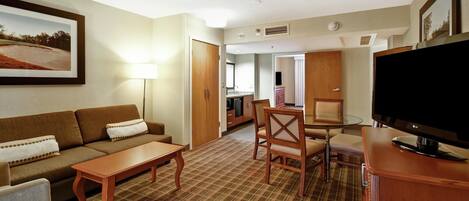 A hotel room with a sofa, a dining table, a TV, and a kitchen area in the background.