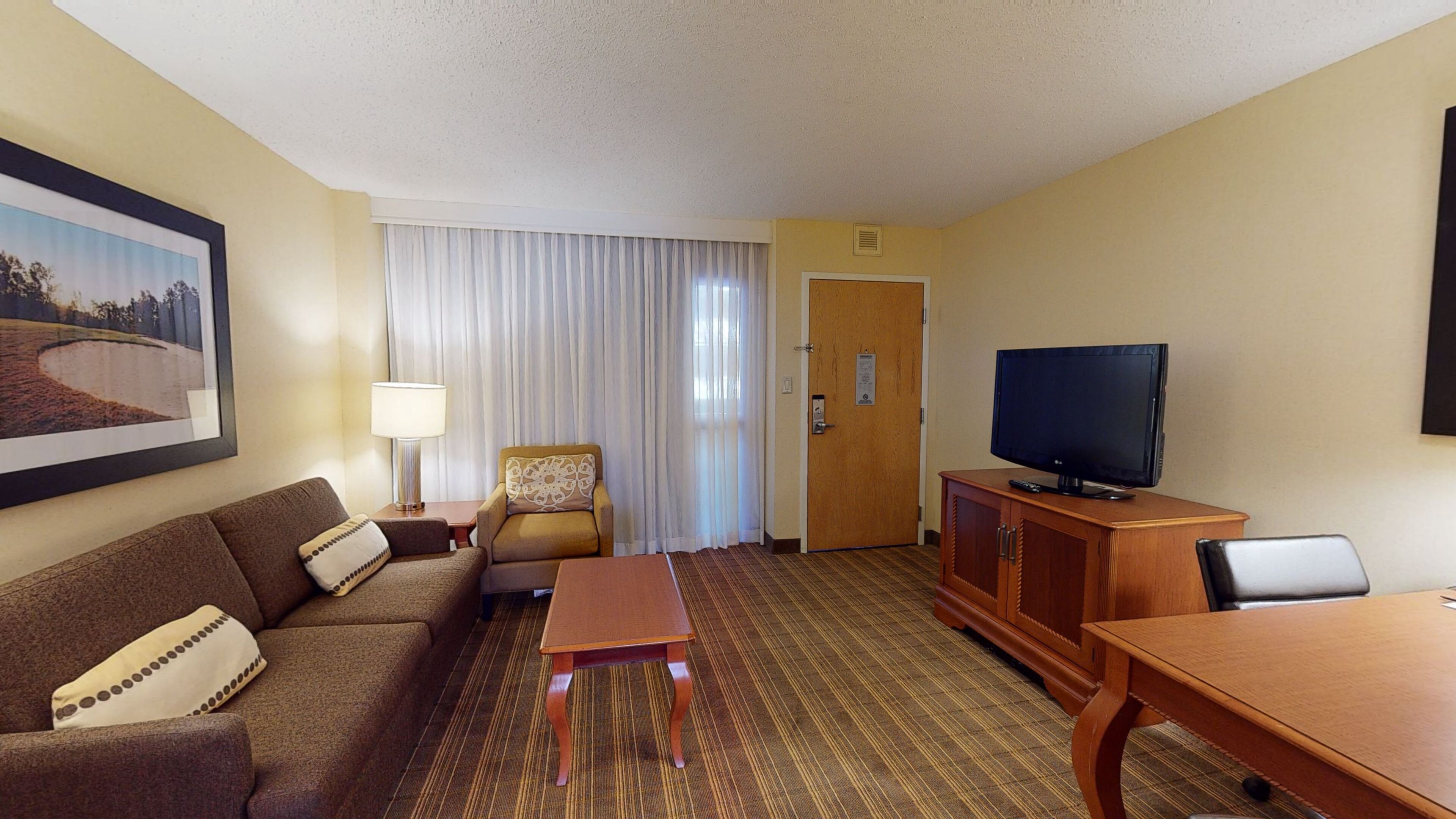 A hotel room with a sofa, armchair, TV, wooden cabinet, and a painting on the wall.