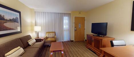A hotel room with a sofa, armchair, TV, wooden cabinet, and a painting on the wall.