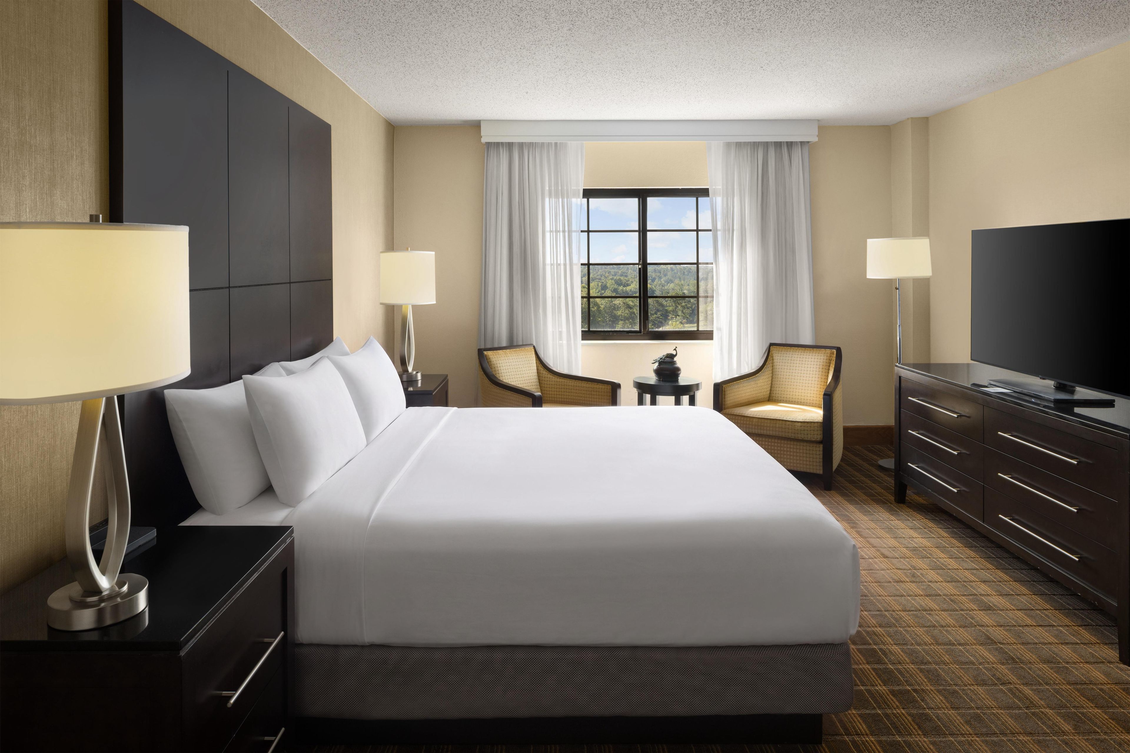 A hotel room with a large bed, two chairs, a TV, and a window with a view.