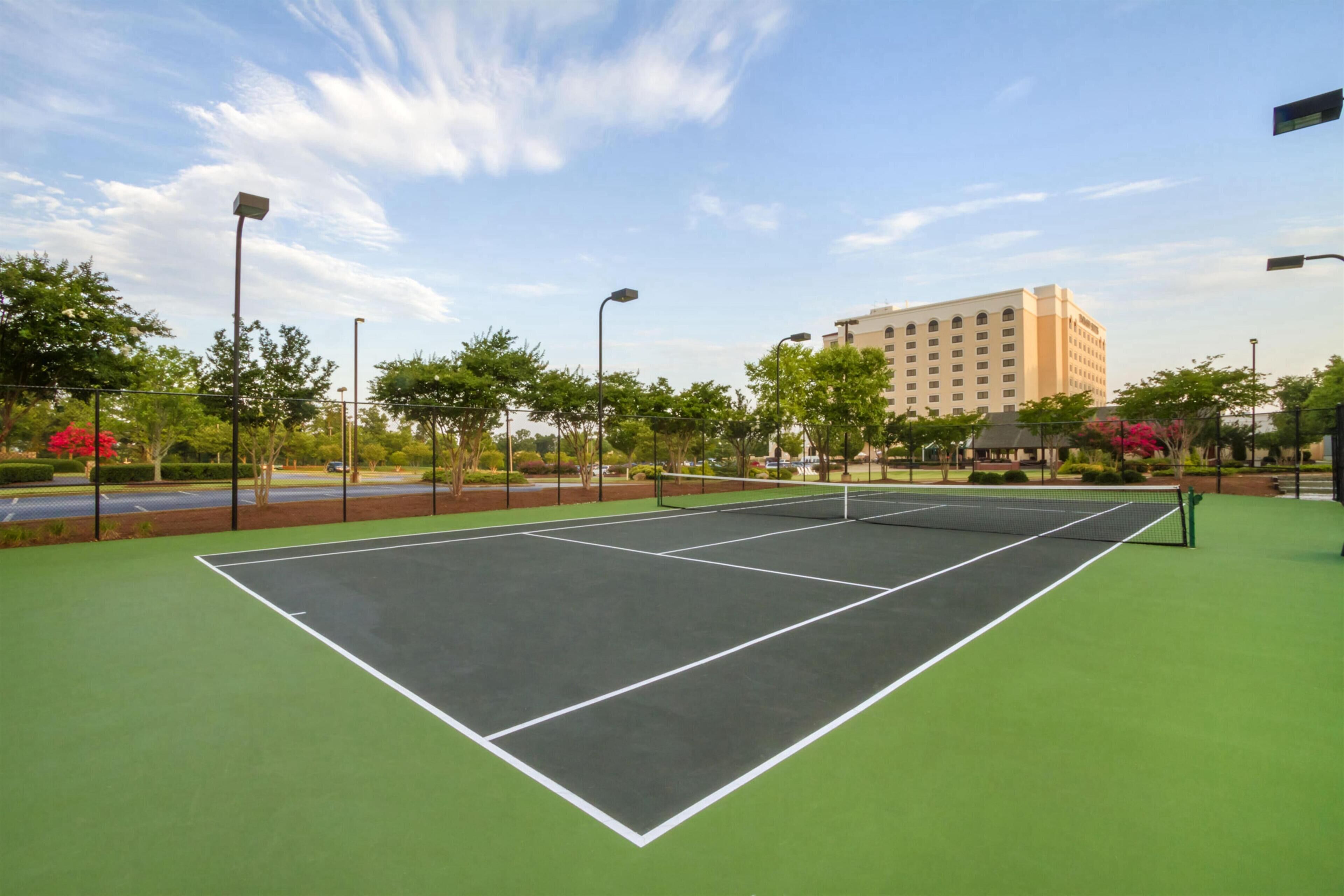 Un court de tennis en plein air, doté d’une surface verte, entouré d’arbres et avec un bâtiment visible à l’arrière-plan.