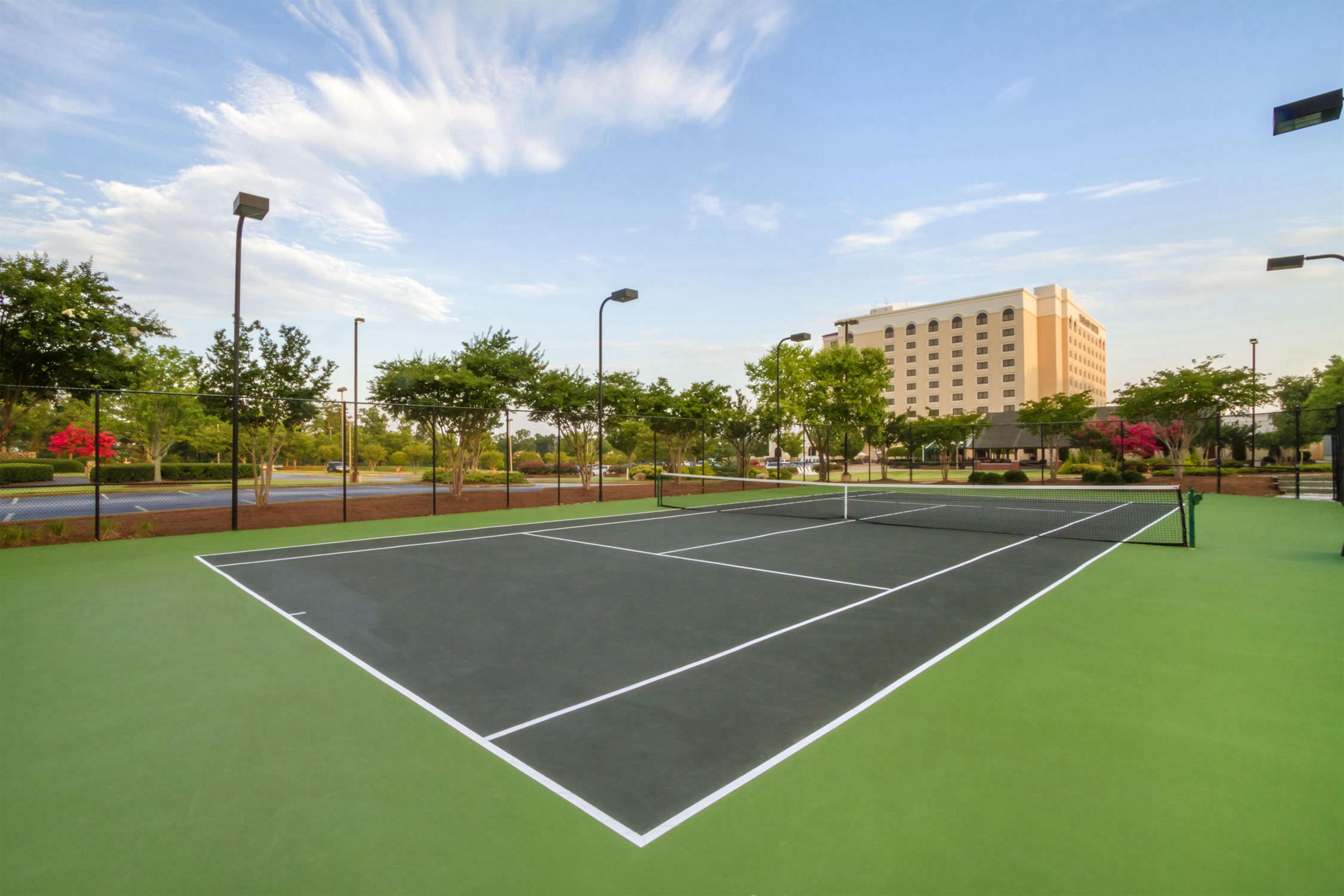 Una cancha de tenis al aire libre con superficie verde, rodeada de árboles y un edificio al fondo.