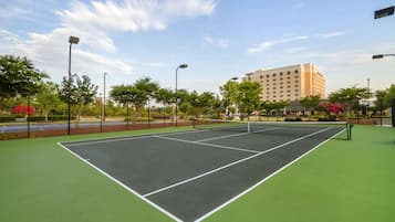 Una cancha de tenis al aire libre con superficie verde, rodeada de árboles y un edificio al fondo.