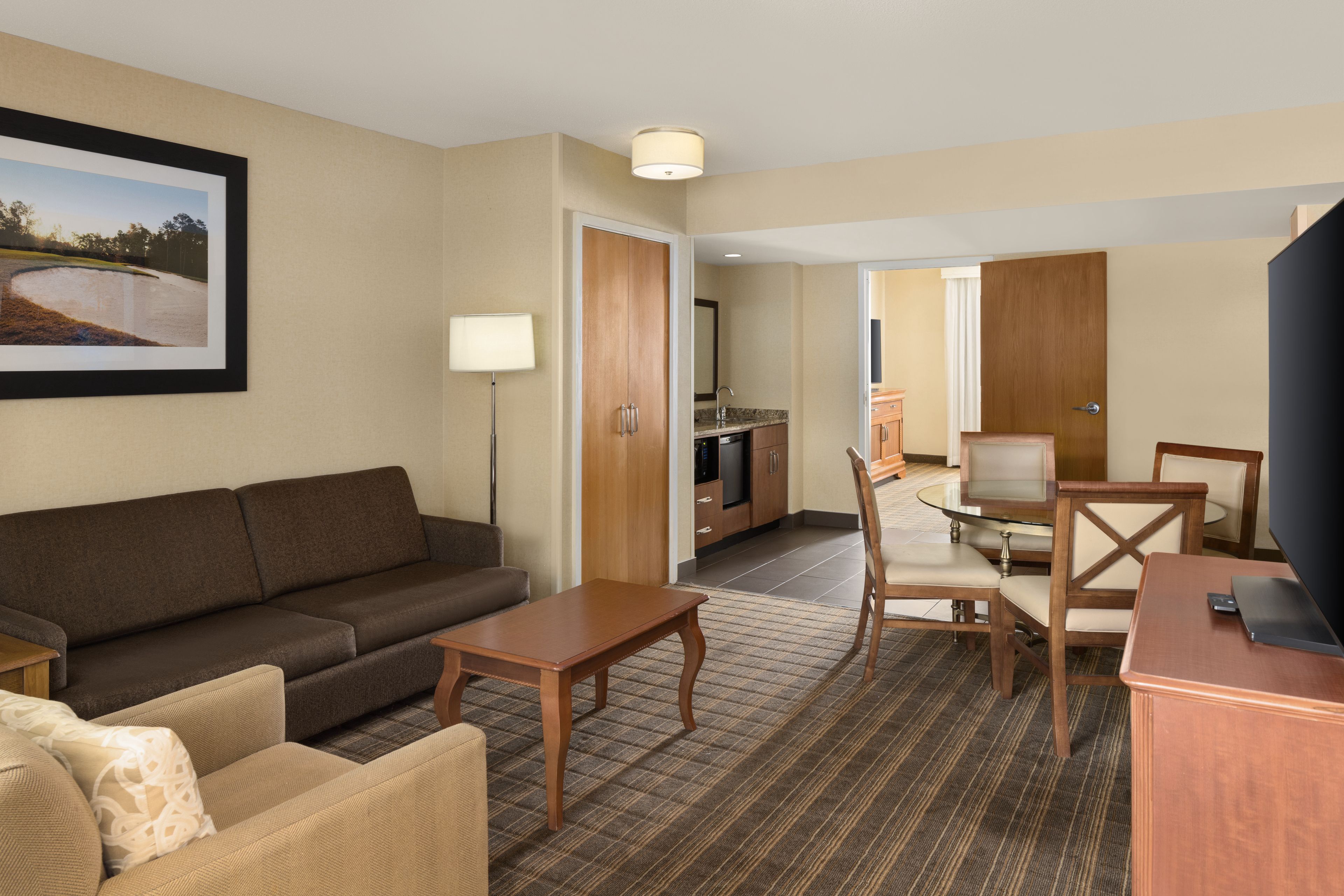 Quarto de hotel com sofá, mesa pequena, televisão, área de refeições com mesa e cadeiras, e cozinha.