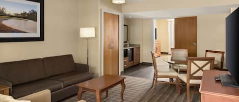 A hotel room with a sofa, a small table, a TV, a dining area with a table and chairs, and a kitchen area.