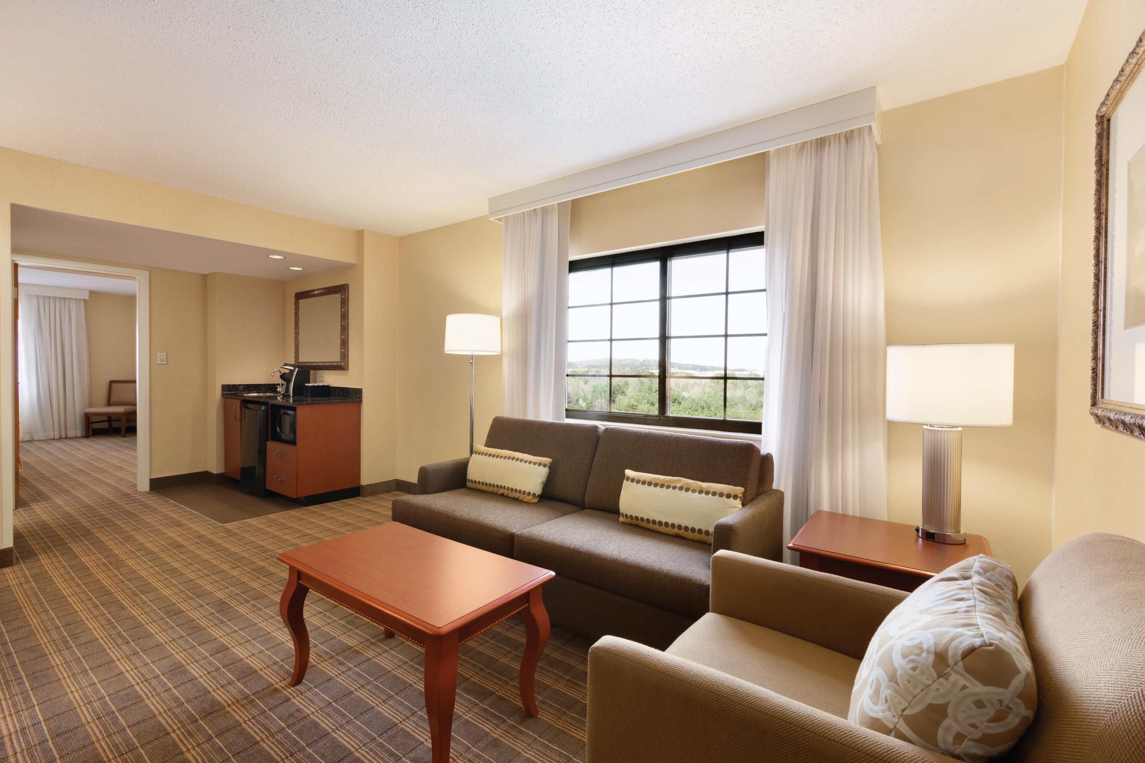 Une chambre d’hôtel équipée d’un canapé, d’un fauteuil, d’une petite table, d’un bureau avec une chaise et d’une grande fenêtre avec des rideaux.