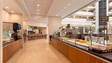 A modern hotel lobby with a buffet area featuring fruit and yogurt, a coffee station, and a view of the upper floors.