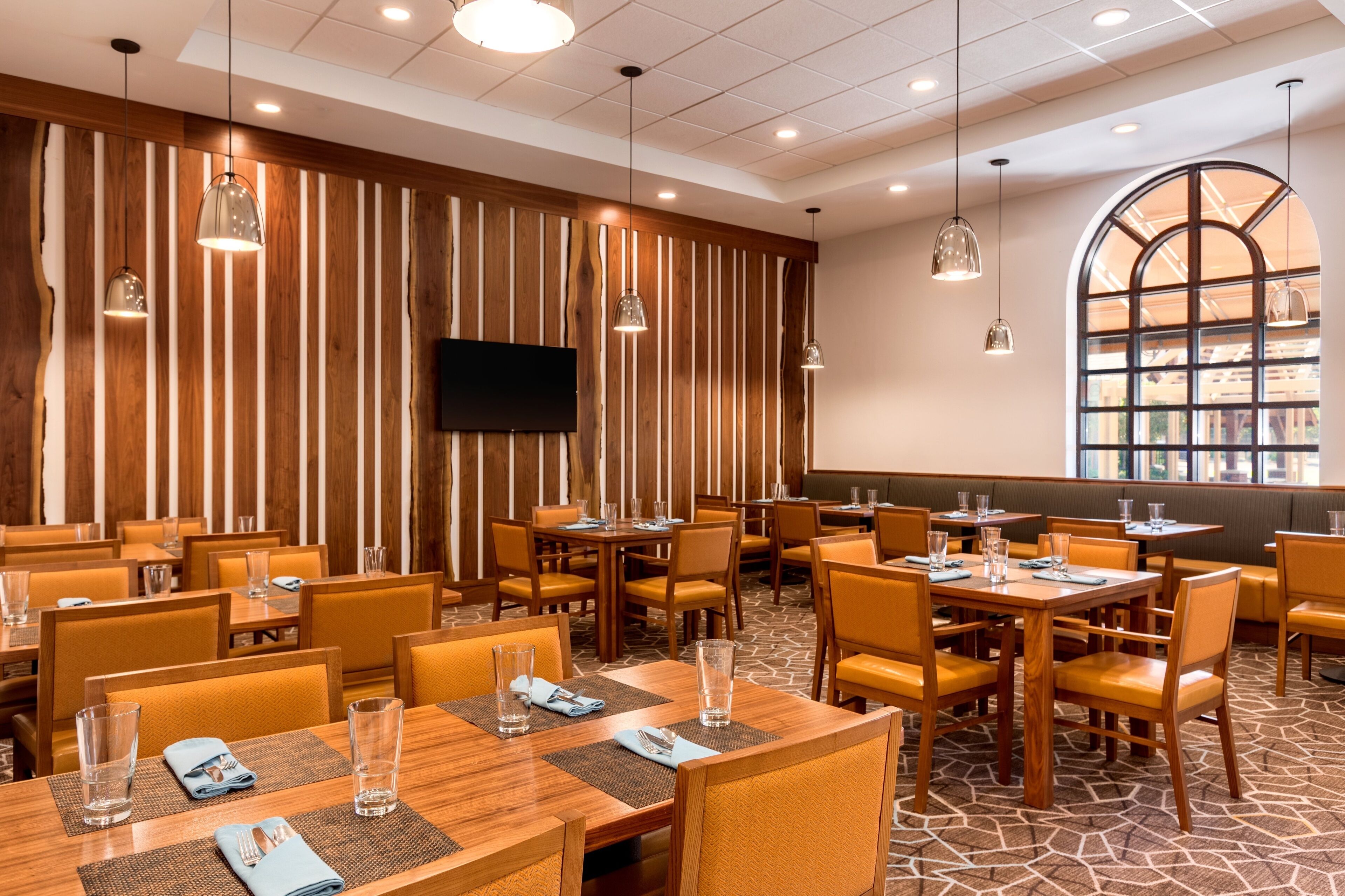 A hotel dining area with wooden panel walls, multiple tables and chairs, hanging pendant lights, and a large arched window.
