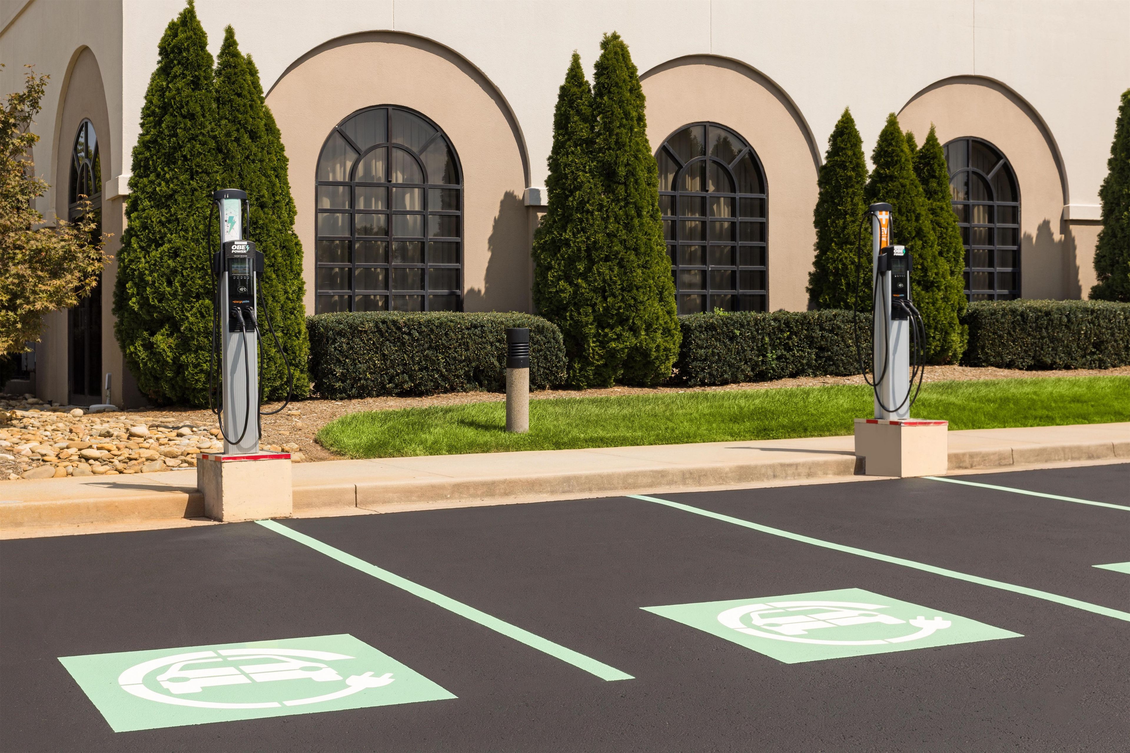 A parking area with electric vehicle charging stations, marked parking spaces, and a building with arched windows and greenery.