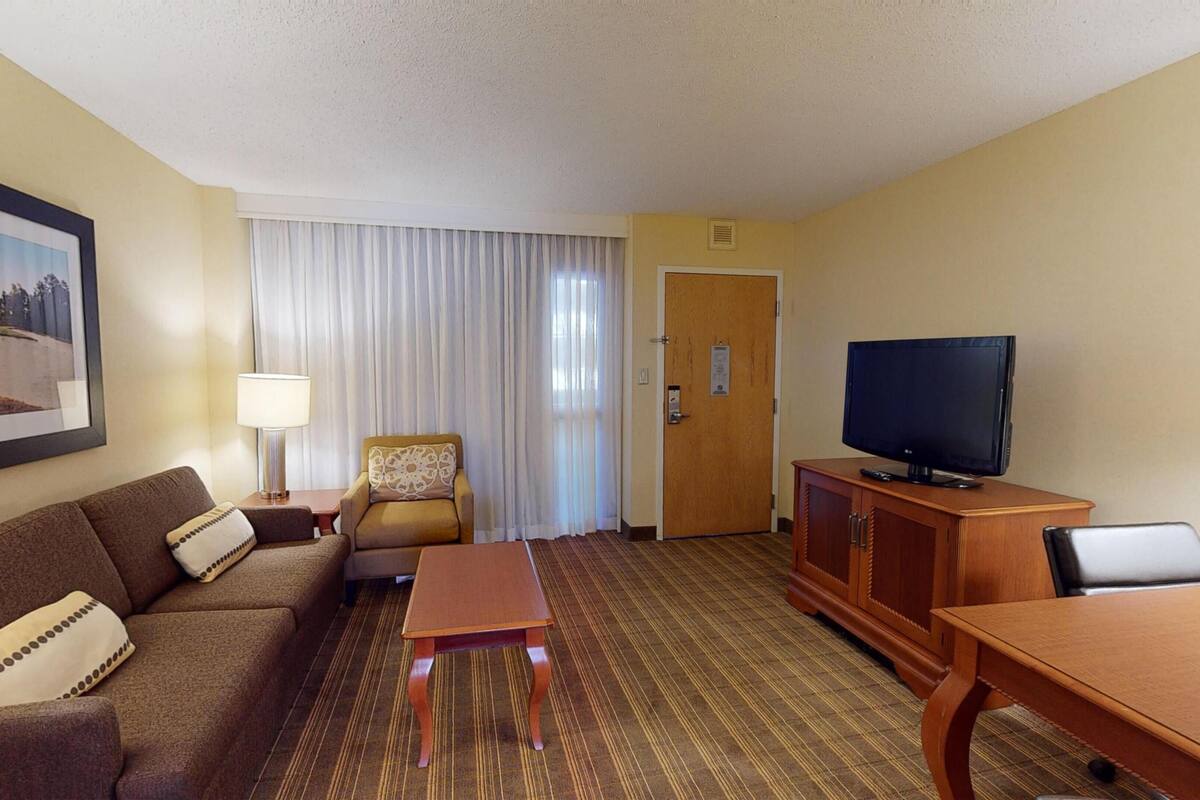 Une chambre d’hôtel équipée d’un canapé, d’un fauteuil, d’une télévision, d’une armoire en bois et d’un tableau accroché au mur.