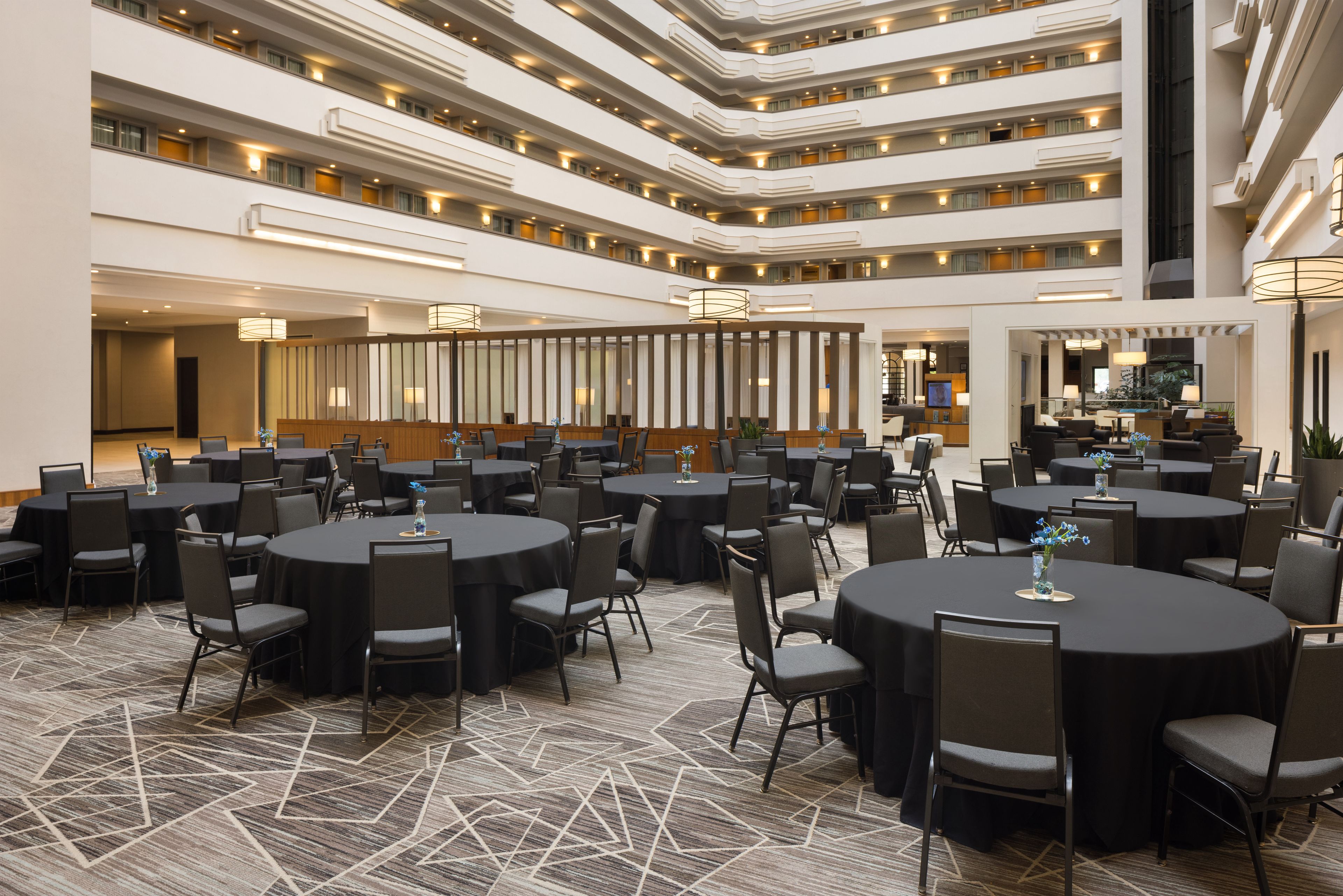 A multi-level hotel lobby with round tables and chairs, a central reception area, and a staircase.