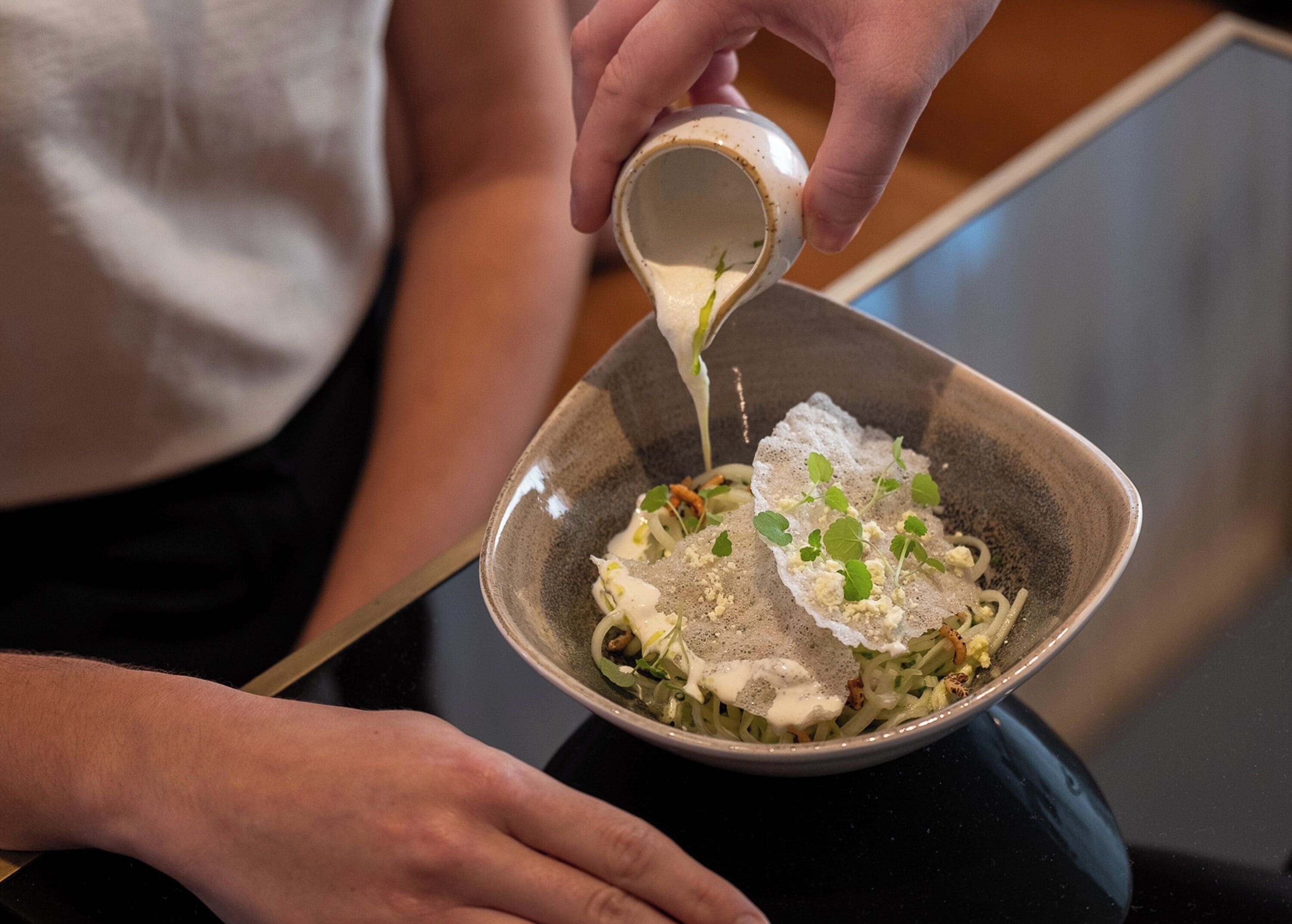 Una persona versa una salsa cremosa in una ciotola con noodles, guarniti con erbe aromatiche e formaggio.