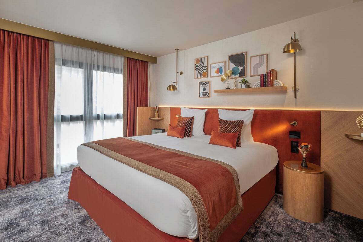 Ein Hotelzimmer mit einem großen Bett, einem Nachttisch, einer Wandablage mit Büchern und einem Fenster mit Vorhängen.