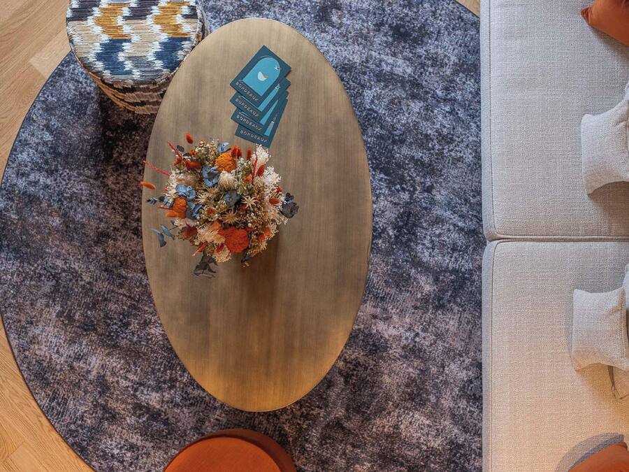 Una habitación con una mesa redonda de madera, una alfombra estampada, un sofá con cojines y un jarrón decorativo con flores.