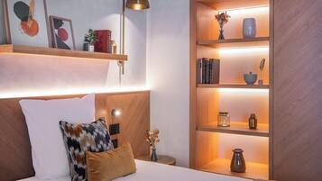 A bedroom with a bed, a nightstand, a shelf with books and decorative items, and a wall-mounted lamp.