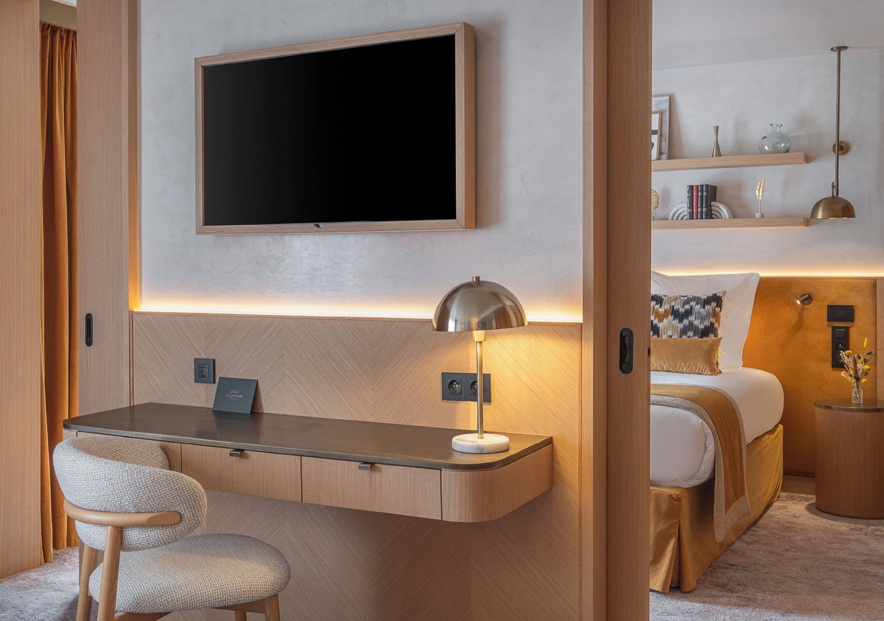 Quarto de hotel moderno com televisão de tela plana fixada acima de uma escrivaninha, luminária de cabeceira, cama com travesseiro estampado e pequena mesa lateral com objetos decorativos.