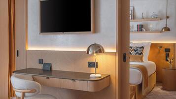 Quarto de hotel moderno com televisão de tela plana fixada acima de uma escrivaninha, luminária de cabeceira, cama com travesseiro estampado e pequena mesa lateral com objetos decorativos.