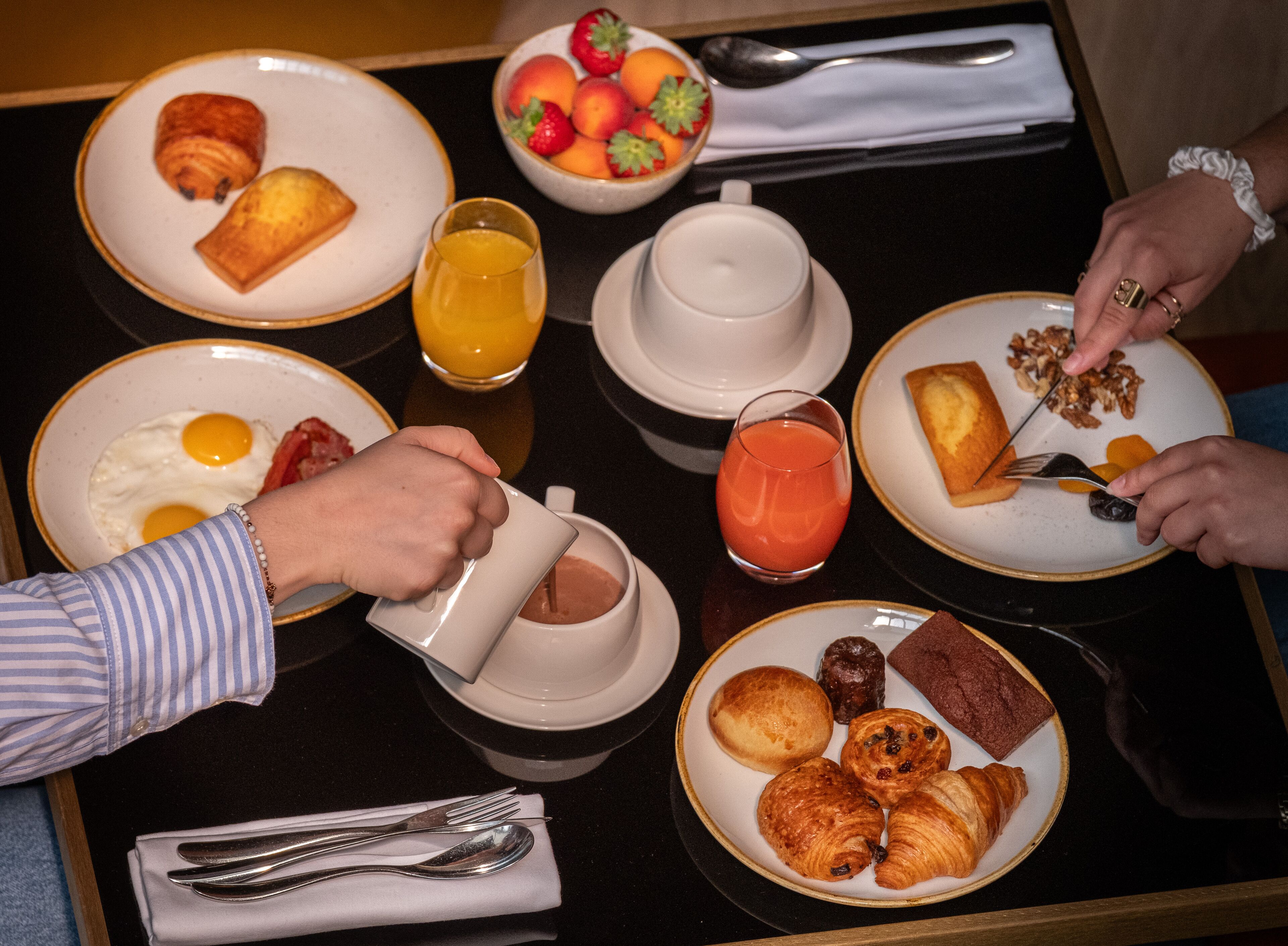 Une table de petit-déjeuner avec des assiettes de nourriture, un saladier de fruits et un verre de jus d’orange.
