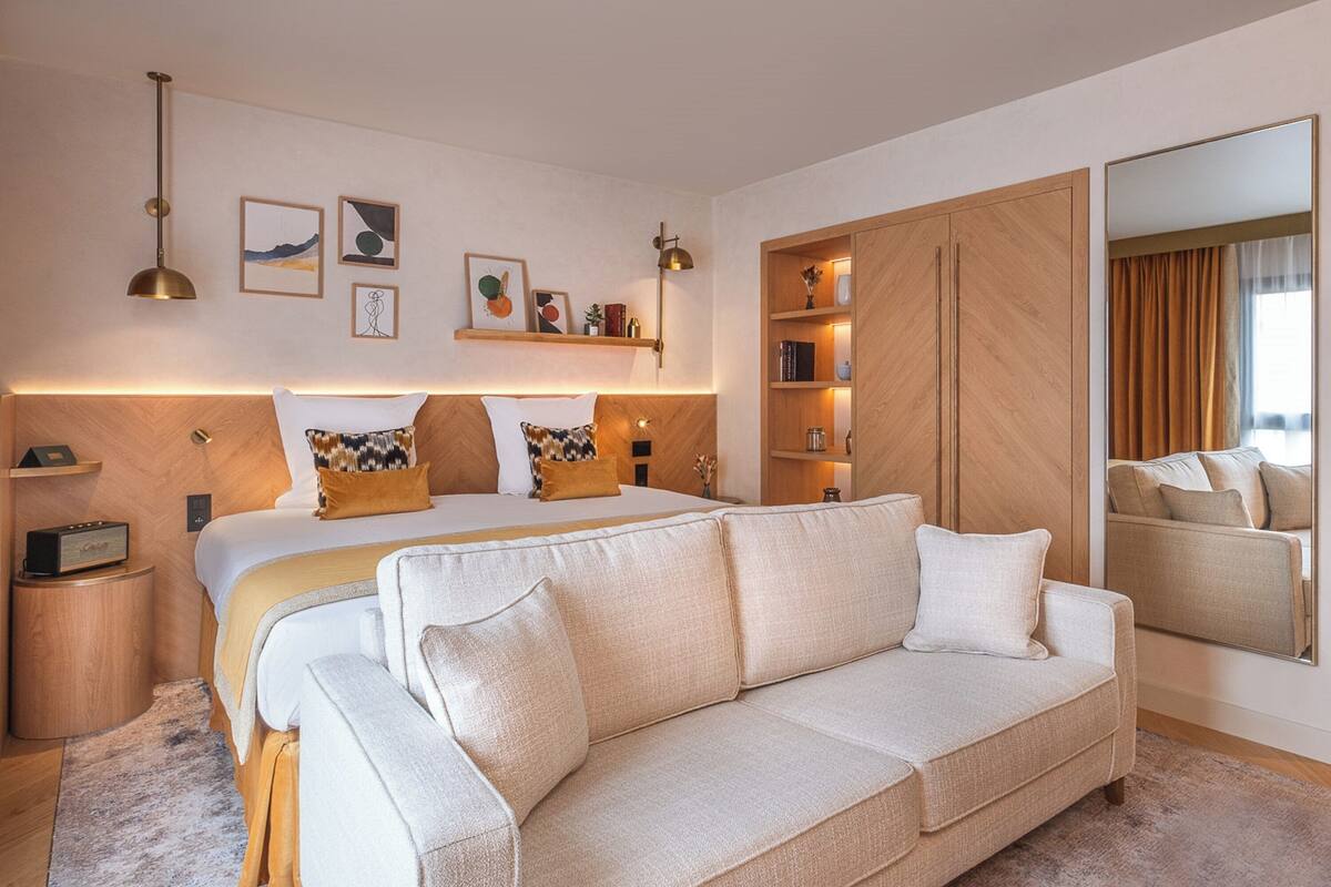Ein modernes Schlafzimmer mit einem großen Bett, einer Couch, einem Nachttisch, einer Lampe, einem Spiegel und Kunstwerken an der Wand.