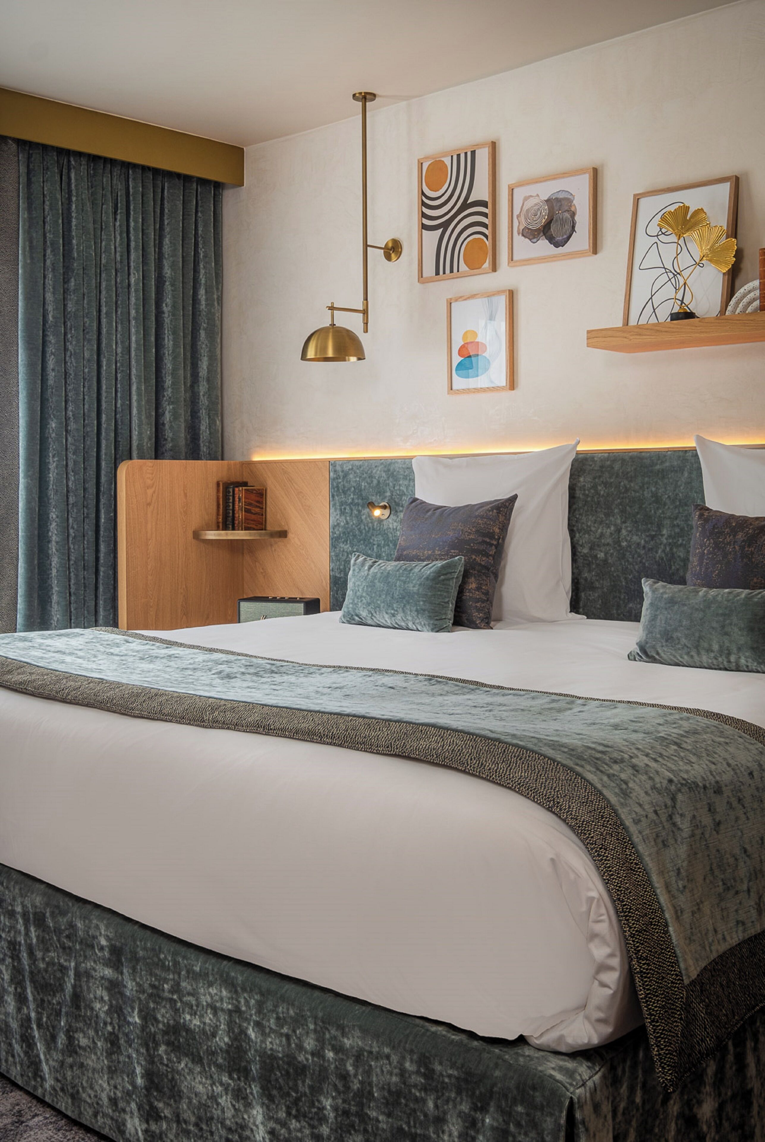 A hotel room with a large bed, a wooden headboard, a bedside table with books, and wall art.