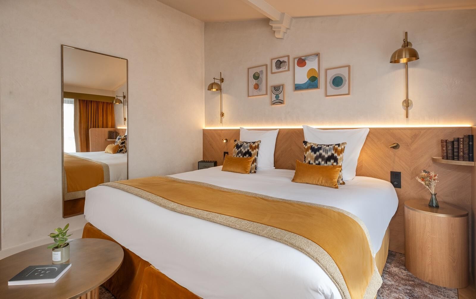 Habitación de hotel con una cama grande, mesitas de noche, un espejo, obras de arte en la pared y una pequeña mesita con un florero.