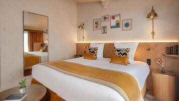 Habitación de hotel con una cama grande, mesitas de noche, un espejo, obras de arte en la pared y una pequeña mesita con un florero.