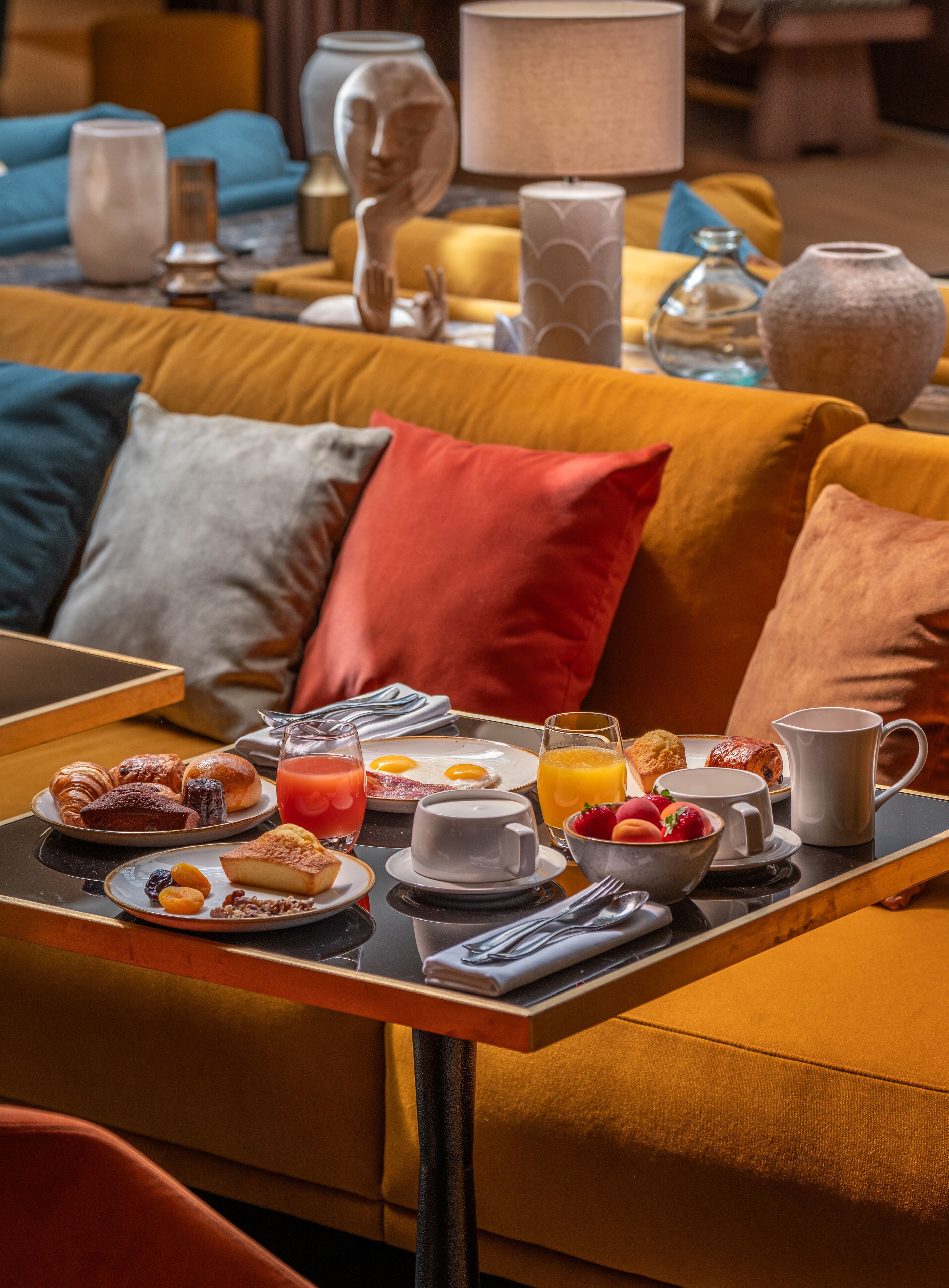 Bandeja de desayuno con pasteles, frutas y bebidas sobre una mesa en un rincón acogedor con un sofá y objetos decorativos.