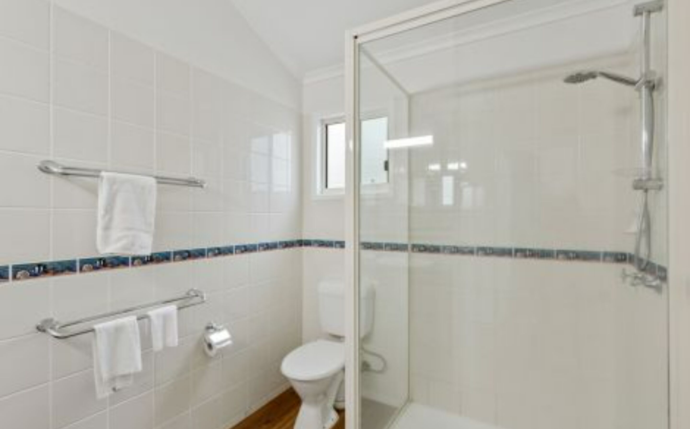 Un baño con azulejos blancos, cabina de ducha de cristal, inodoro y toallero.