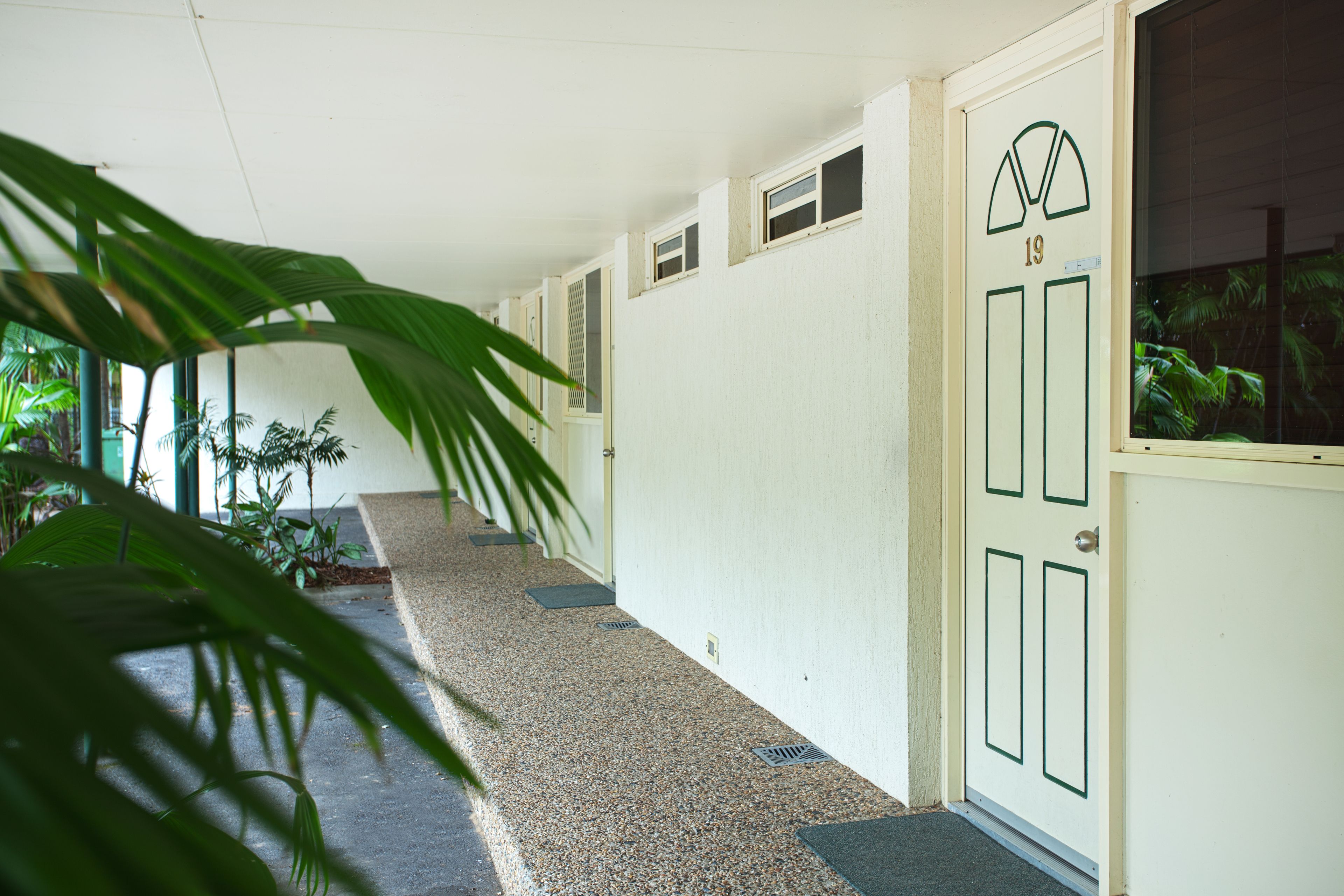 Un pasillo con una puerta marcada '19', piso de baldosas y plantas en macetas a lo largo de la pared.