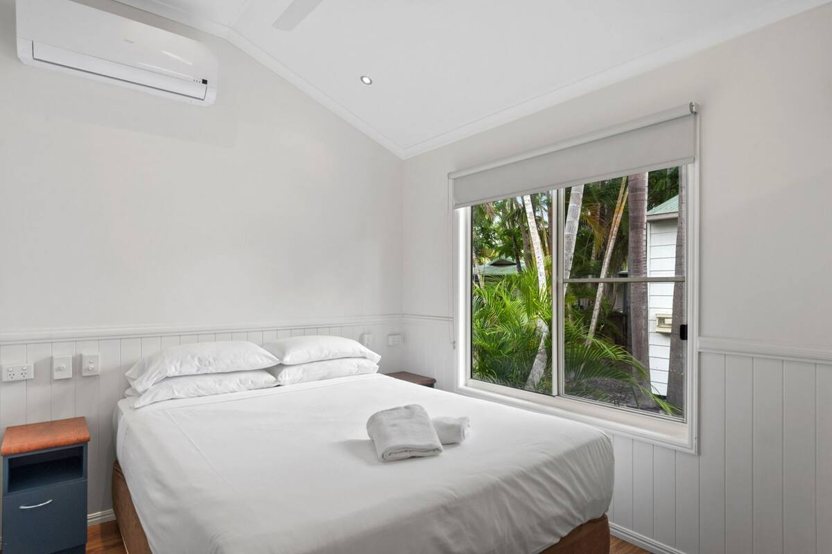 A bedroom with a bed, white walls, a window with a view of greenery, and an air conditioning unit.